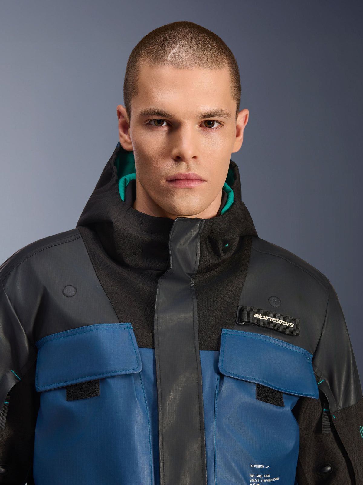 Alpinestars Mospher Waterproof Jacket, Black and Dark Petrol, close-up of male model wearing the utility-inspired parka, featuring dark blue chest pockets, black hood with teal lining, and Alpinestars branding on the chest.