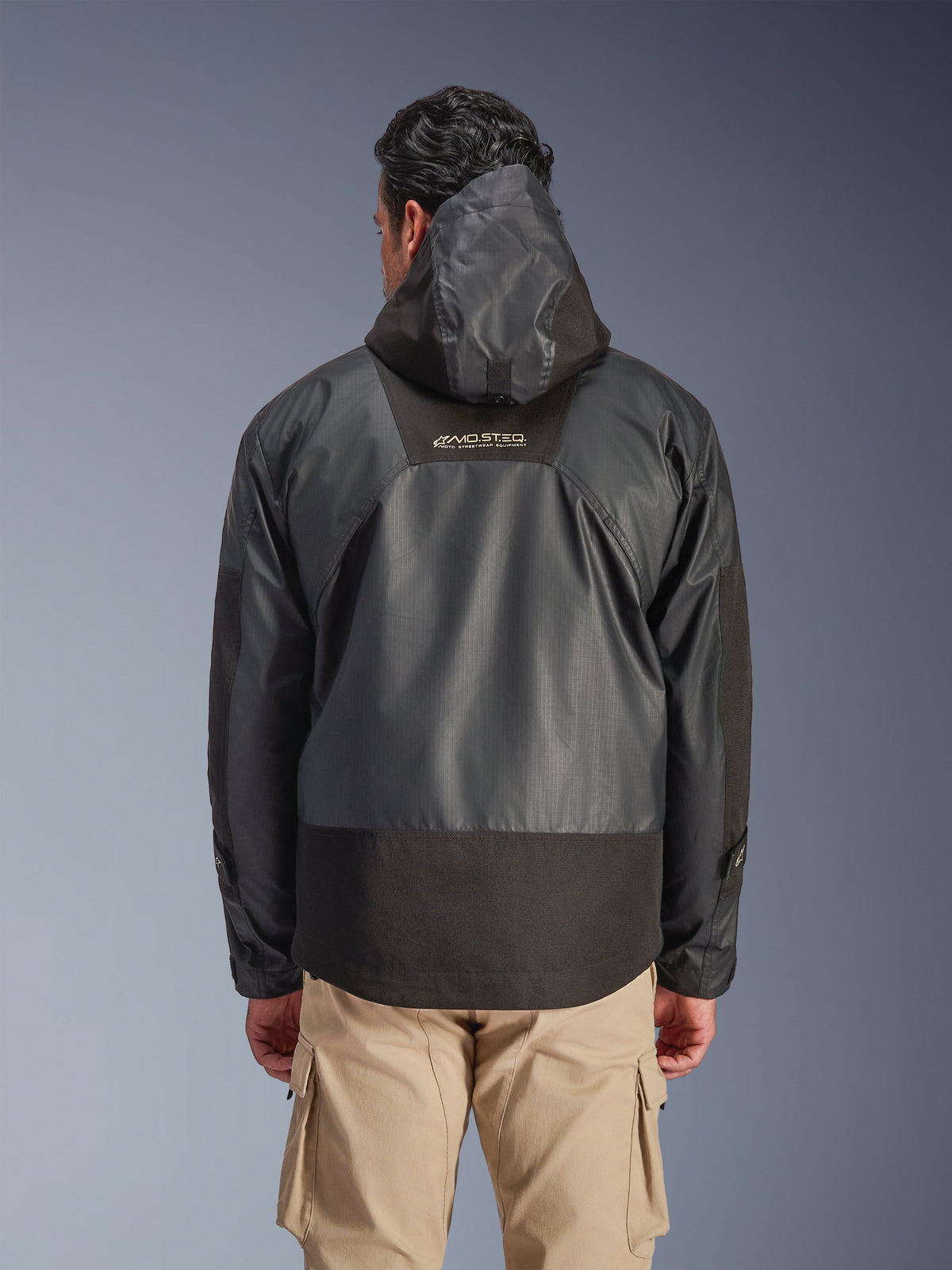 Alpinestars Mospher Waterproof Jacket i svart, vy bakifrån på en man, med huva och slitstark ripstop-textilkonstruktion, visas med beige cargobyxor, framhäver MO.ST.EQ.-logotypen på övre ryggen, urban motorcykeldesign.