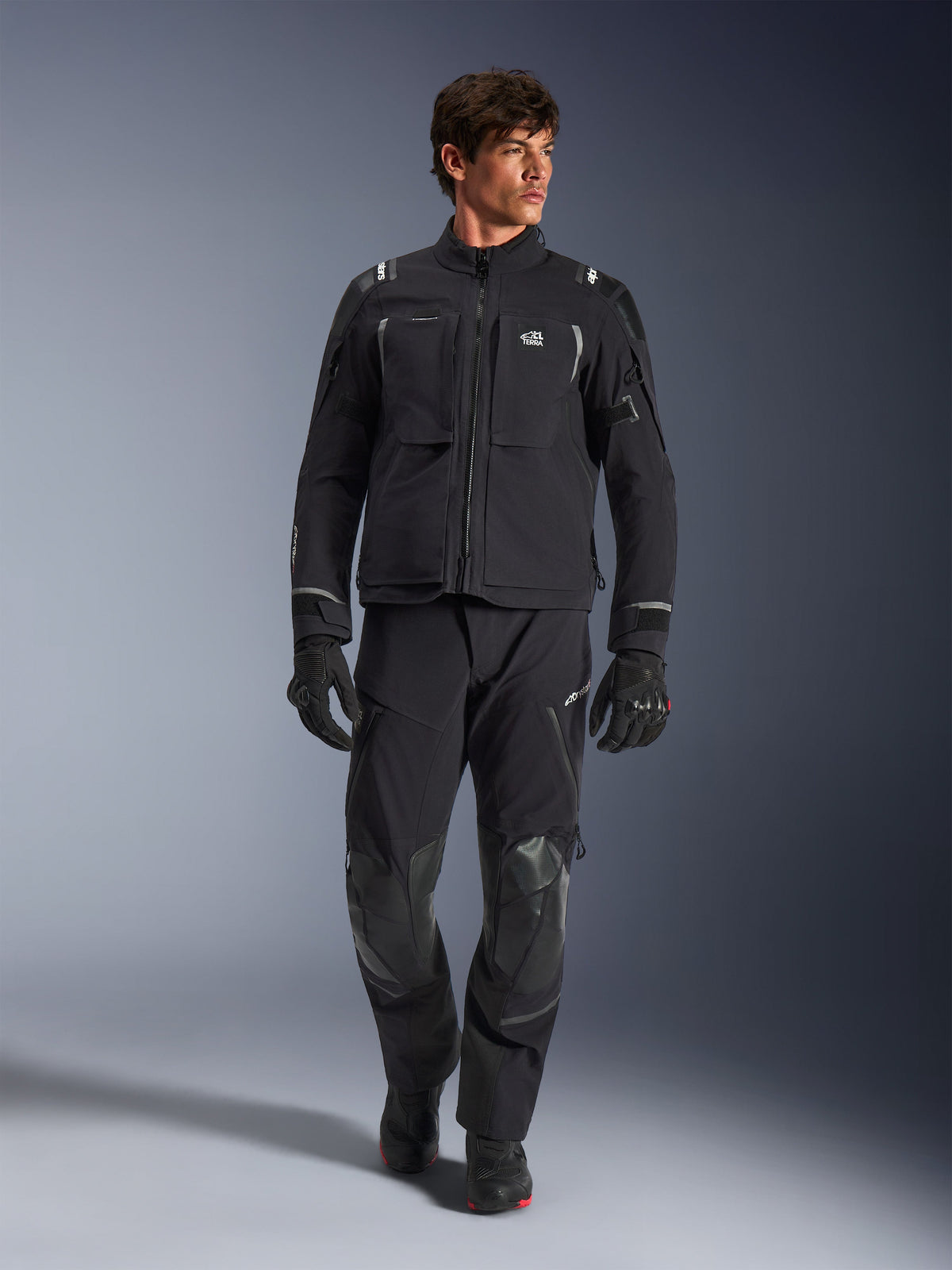Male model wearing Alpinestars Andes Pro Drystar® XF Jacket, Black and Dark Gray adventure motorcycle jacket, paired with matching technical pants and gloves, featuring multiple pockets and 'Terra' branding on the chest.