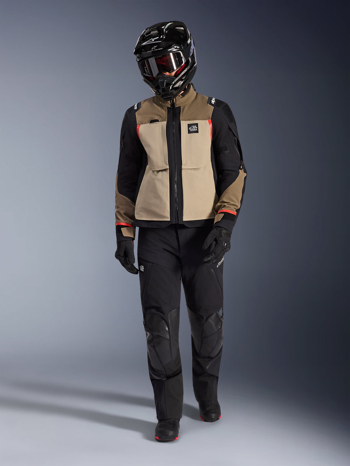 Alpinestars Andes Pro Drystar® XF-jacka i Walnut Aluminum, bärs med svarta adventure-byxor, svarta handskar och en svart helhjälm med goggles, med röda detaljer och All Terra-logotyp, professionell adventure touring-utrustning