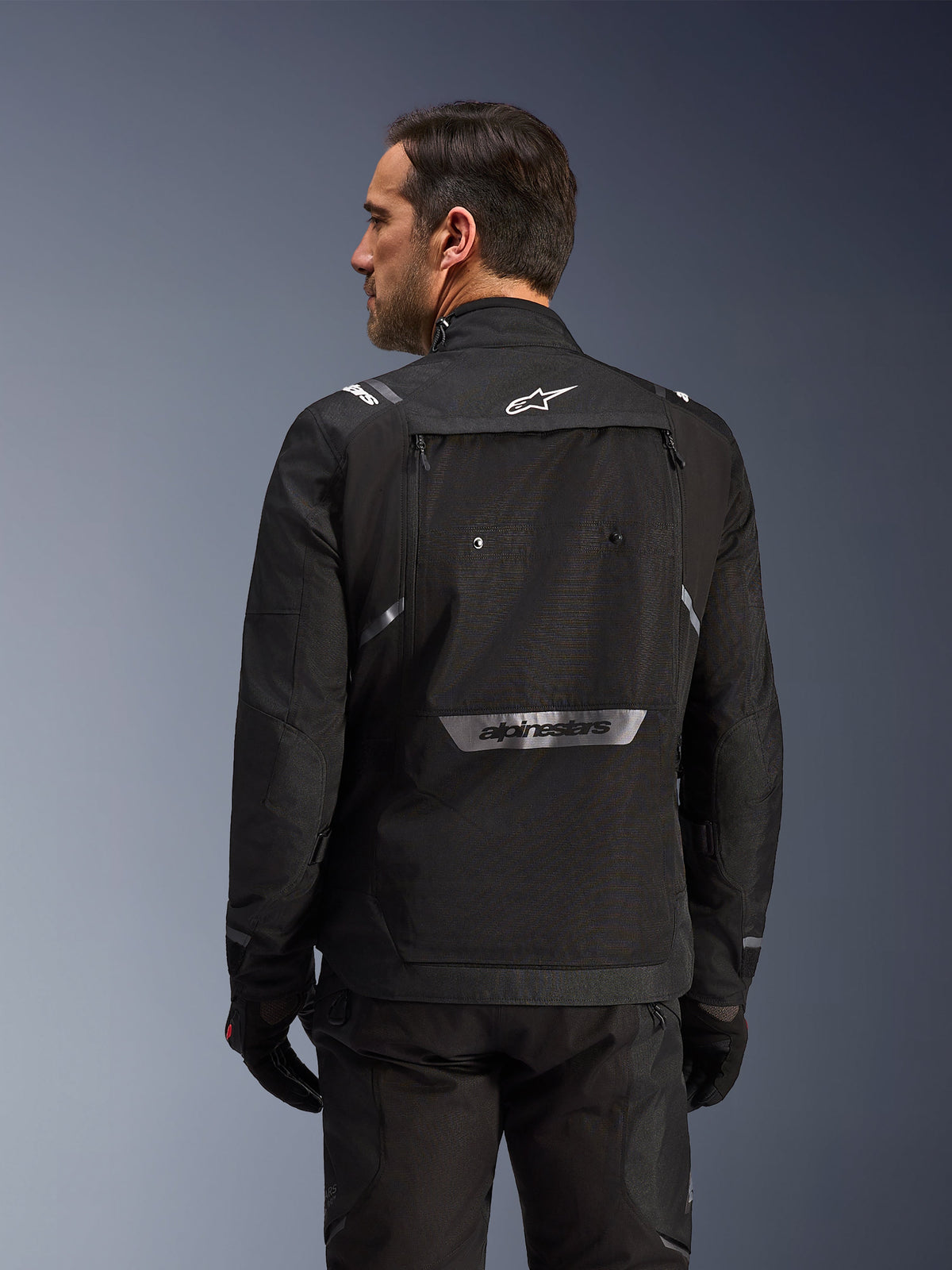 Motorcyklist sedd bakifrån som bär Alpinestars Cusco Drystar®-jacka i svart, äventyrstouringjacka i textil med stor förvaringsficka på ryggen och reflekterande logotyp, matchad med svarta byxor och handskar