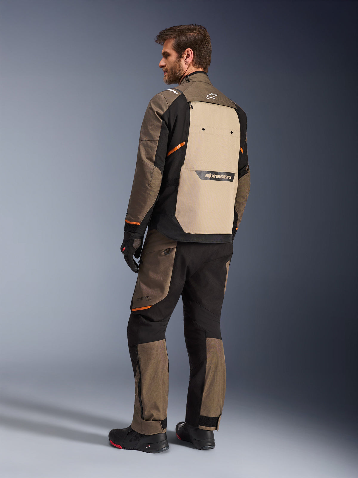 Alpinestars Cusco Drystar®-jacka i Walnut Aluminum Black, vy bakifrån som visar beige ryggpanel och orange detaljer, bärs med matchande motorcykelbyxor, svarta handskar och stövlar, professionell adventure touring-utrustning