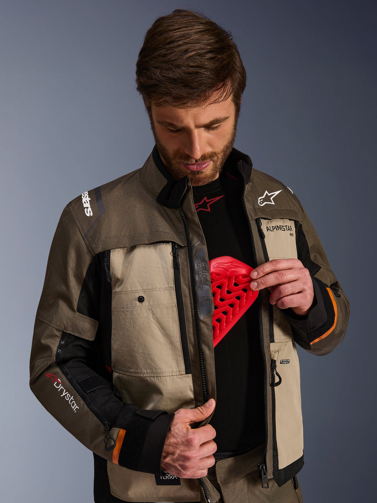 Alpinestars Cusco Drystar®-jacka, Walnut Aluminum Black, bärs av en man som demonstrerar bröstskyddsfickan med ett rött skyddsinlägg, textilmotorcykeljacka med ventilationsöppningar med dragkedja och märkesdetaljer för adventure touring.