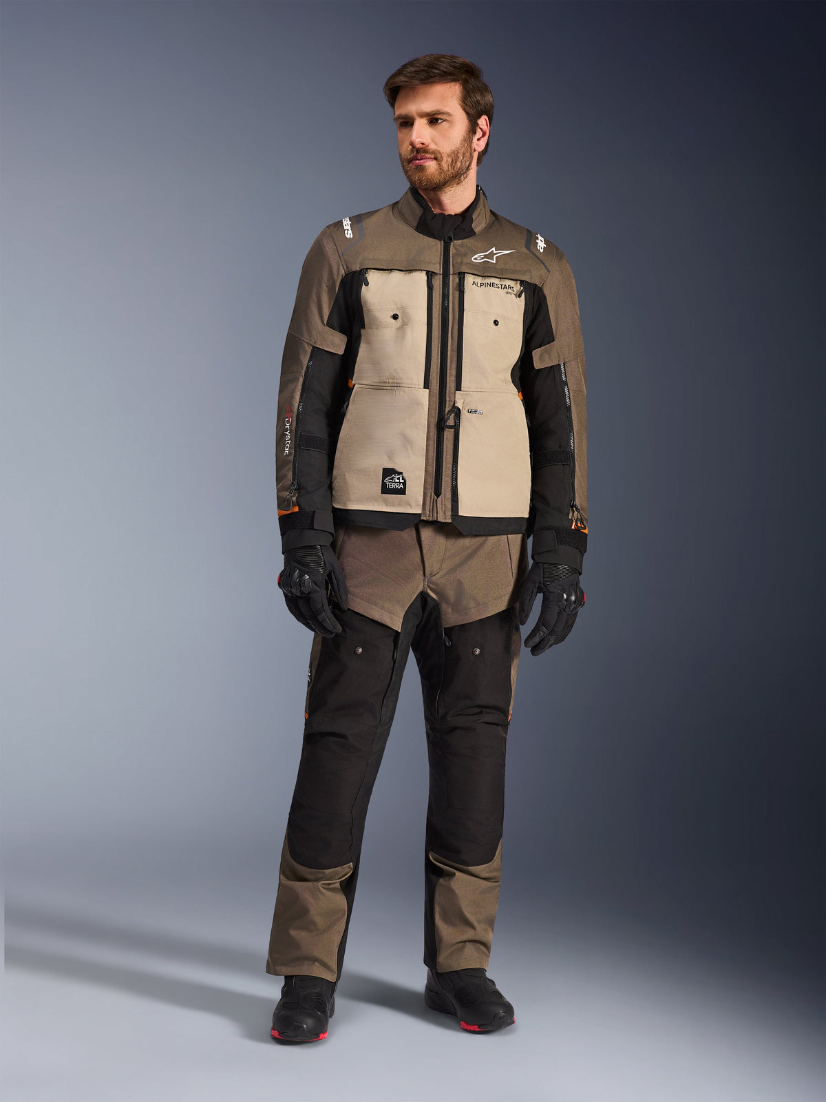 Alpinestars Cusco Drystar®-jacka i Walnut Aluminum Black, bärs av en manlig förare med matchande adventure touring-byxor, svarta handskar och stövlar, med beigea bröstpaneler, svarta ärmar med subtila orangea detaljer och funktionella ventilationsdragkedjor för landsvägskörning.