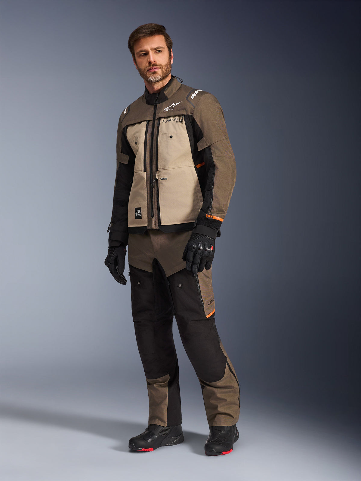 Alpinestars Cusco Drystar®-jacka i Walnut Aluminum Black, bärs av en manlig modell med matchande motorcykelbyxor och handskar, adventure-touring-design med beiga bröstpaneler och svarta detaljer.