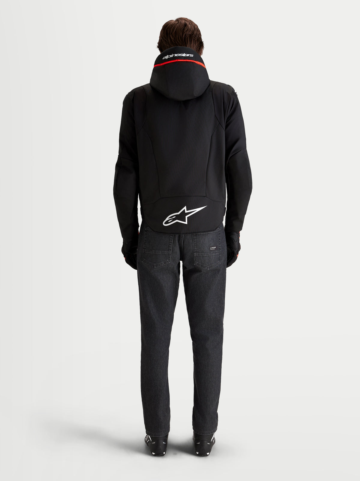 Alpinestars Zaca V2 Waterproof Jacka, vattentät motorcykeljacka för herr, svart, vy bakifrån med integrerad huva, förlängd bakre fåll med vit Astar-logotyp och mörka jeans