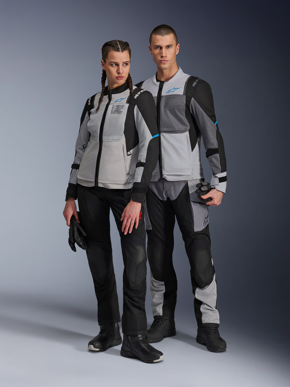 Man och kvinna i Alpinestars Stella ST-2 Air-jackor och matchande tekniska byxor, färgställningen Ice Gray och Black med blå detaljer, mesh-ventilation och 'ALPINESTARS EXPLORATION EQUIPMENT'-märkning, studiobild