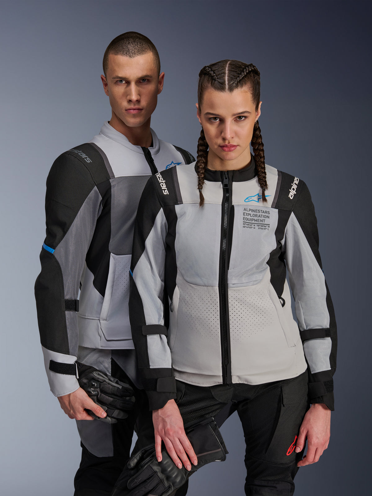Alpinestars ST-2 Air- och Stella ST-2 Air-jackor, isgrå och svarta, textila motorcykeljackor med meshpaneler och laserperforeringar, med 'Alpinestars Exploration Equipment'-märkning, som bärs av en manlig och en kvinnlig modell som håller i mc-handskar.