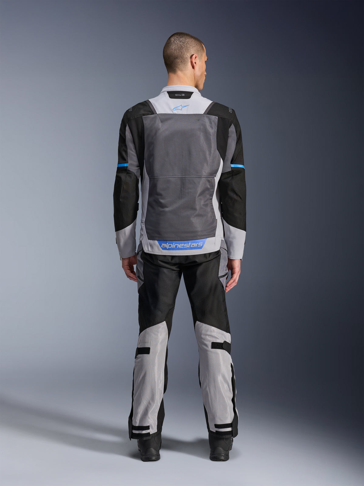 Alpinestars ST-2 Air-jacka, Ice Gray Dark Gray Black, vy bakifrån på modell, med stora ventilationspaneler i mesh, blå detaljer och Alpinestars-logotyp, kombinerad med matchande motorcykelbyxor för adventure touring.