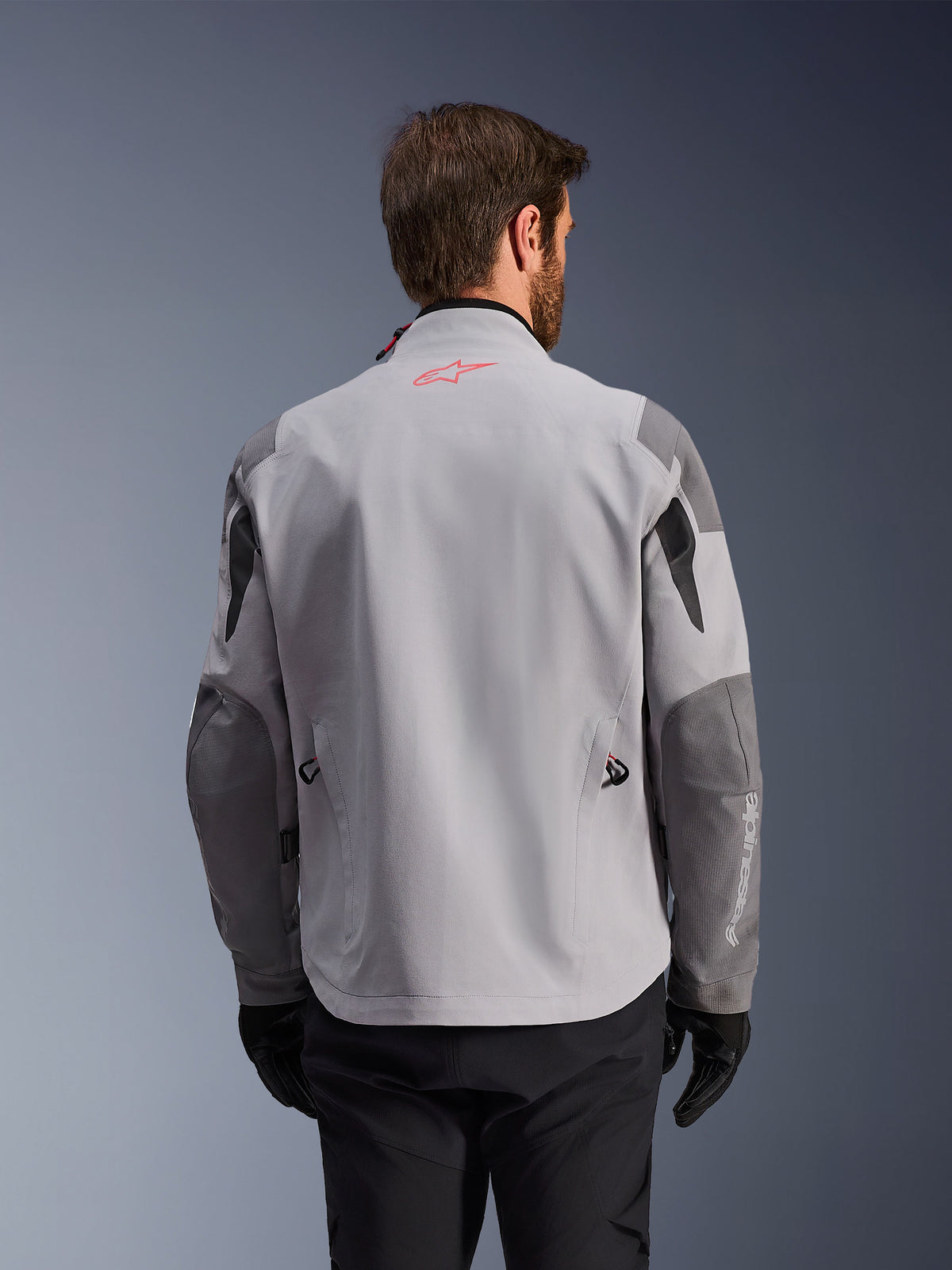Alpinestars Maxdura Drystar®XF Laminated Armored Shell, baksida av äventyrsmotorcykeljacka, mellangrå och mörkgrå, med en röd stjärnlogotyp på kragen och vit vertikal logotyp på ärmen, bärs med svarta körhandskar och byxor