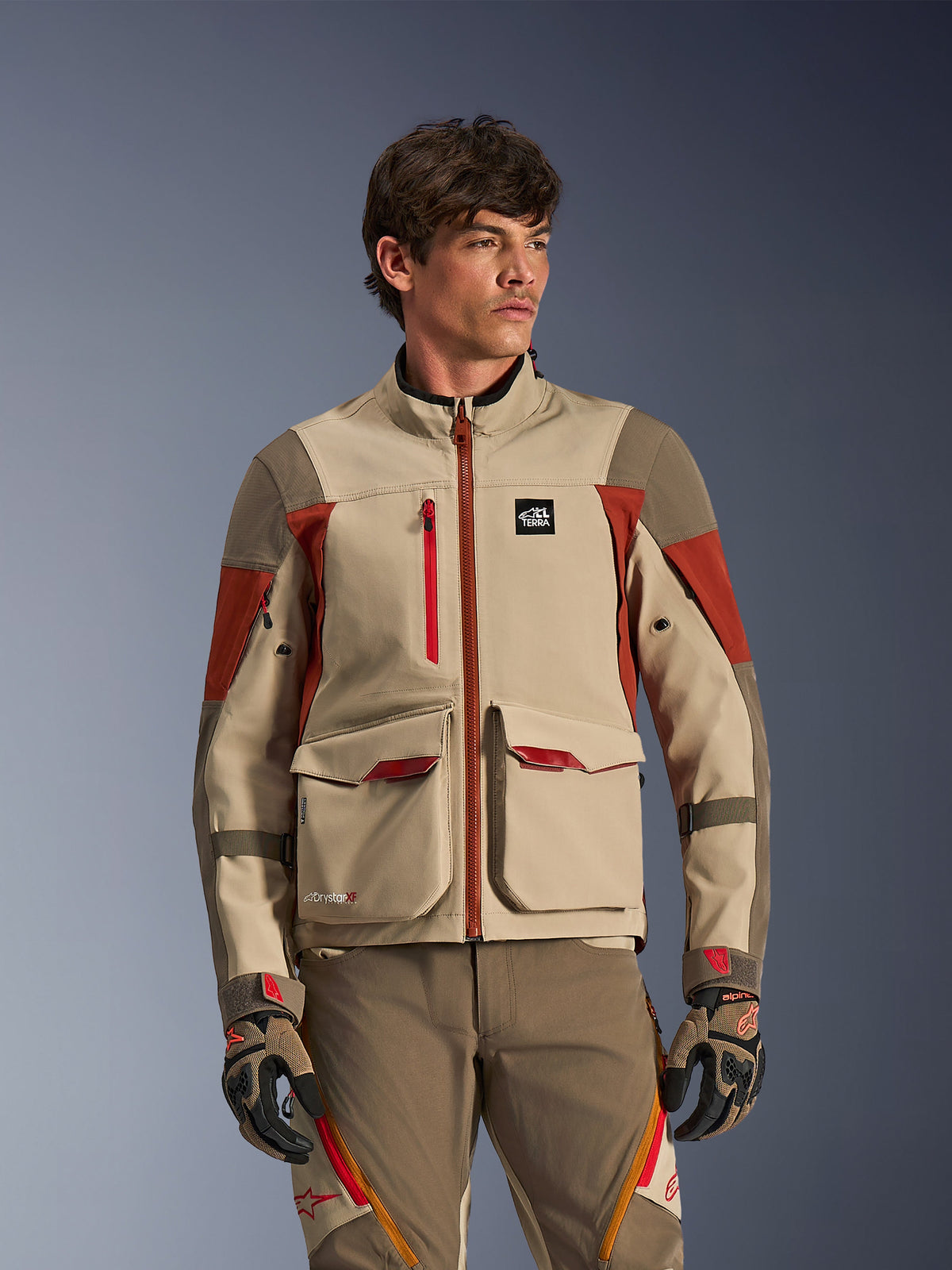 Alpinestars Maxdura Drystar®XF Laminated Armored Shell, Aluminum Walnut Rust Brown, äventyrsmotorcykeljacka buren av en manlig modell, med stora utanpåfickor framtill och tekniska röda detaljer, kombinerad med matchande äventyrsbyxor och handskar.