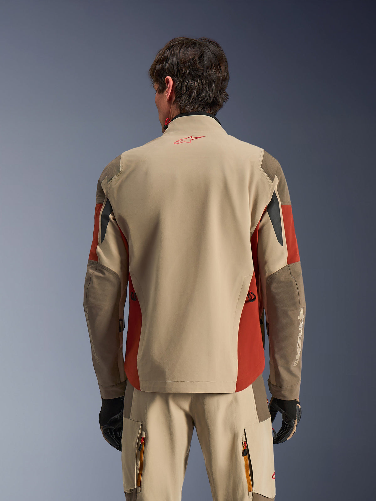 Man som bär Alpinestars Maxdura Drystar®XF Laminated Armored Shell bakifrån, äventyrsjacka i Aluminum Walnut och Rust Brown, matchande tekniska byxor och svarta handskar, 3-lagers laminerad textilkonstruktion med förstärkta paneler, koordinerad outfit för motorcykel-touring