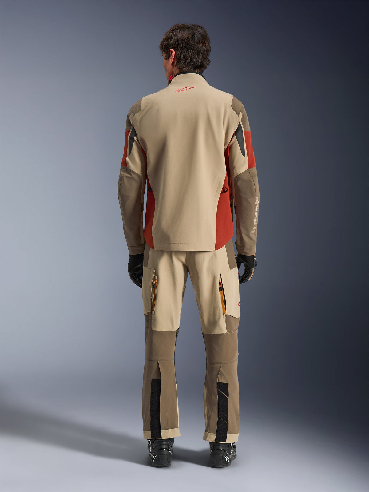 Vy bakifrån av en förare som bär Alpinestars Maxdura Drystar®XF Laminated Armored Shell och matchande äventyrsbyxor i Aluminum Walnut Rust Brown, beige och brunt textilmaterial med orangea detaljer, kombinerat med svarta motorcykelstövlar och handskar, professionell utrustning för äventyrstouring.