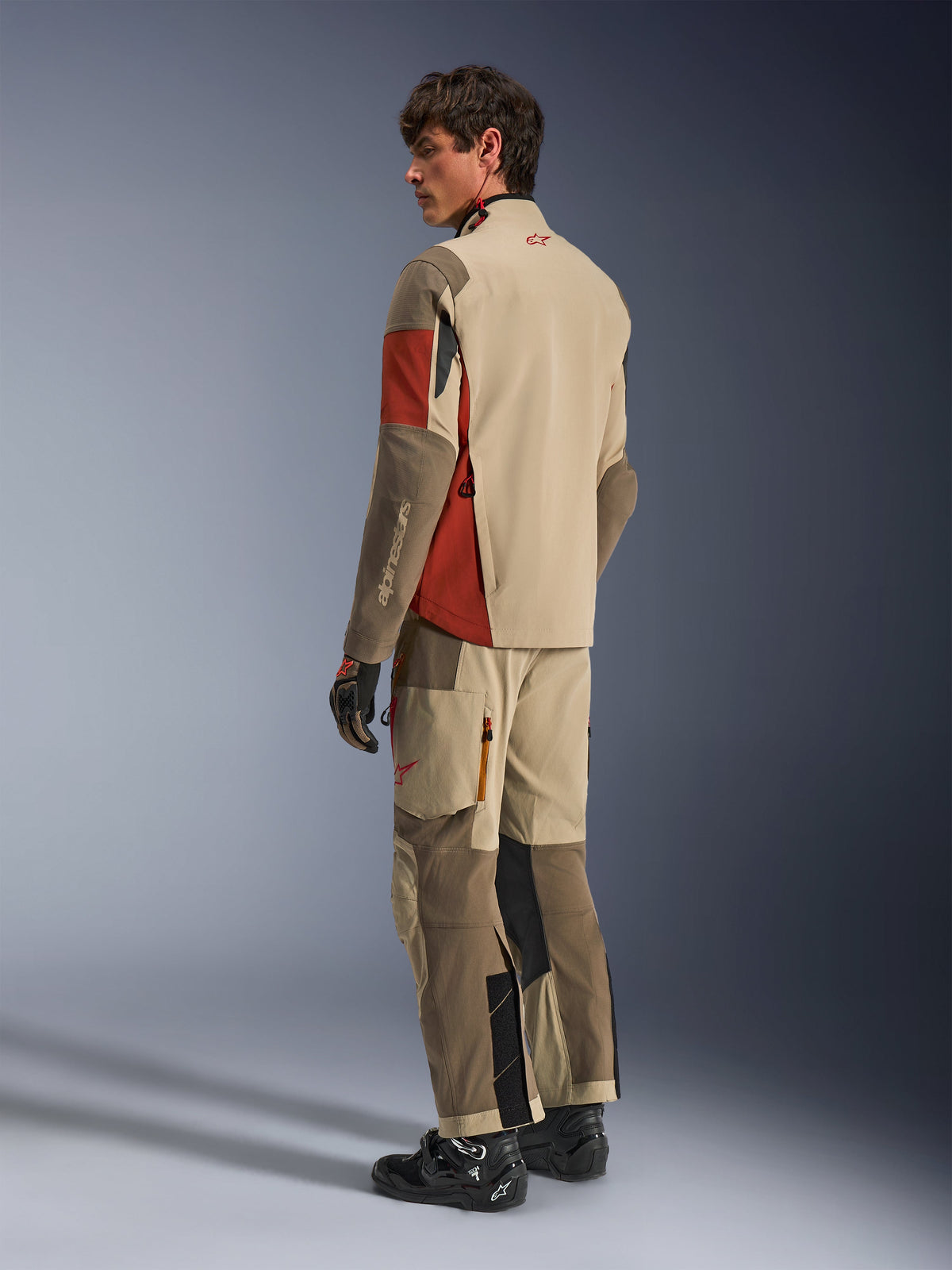 Manlig modell iklädd Alpinestars Maxdura Drystar®XF Laminated Armored Shell i Aluminum Walnut Rust Brown, matchande adventure touring-byxor, svarta handskar och svarta stövlar, vy bakifrån som visar den tekniska textilkonstruktionen och förstärkta zoner.