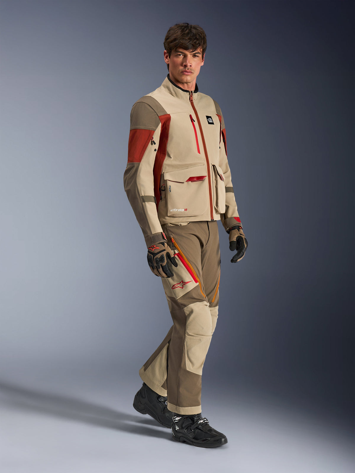 Manlig modell bär Alpinestars Maxdura Drystar®XF Laminated Armored Shell, äventyrsjacka i Aluminum Walnut Rust Brown med matchande byxor, svarta och bruna handskar samt svarta stövlar, teknisk design i flera lager med cargo-fickor och röda dragkedjedetaljer, koordinerad outfit för äventyrstouring
