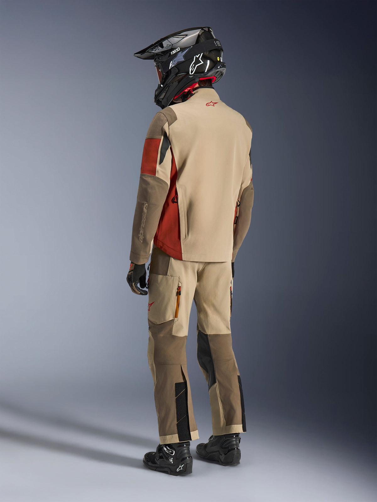 Äventyrsförare sedd bakifrån bärande Alpinestars Maxdura Drystar®XF Laminated Armored Shell i Aluminum Walnut Rust Brown, tillsammans med matchande körbyxor, svart off-road-hjälm och svarta stövlar, komplett teknisk touring-outfit.