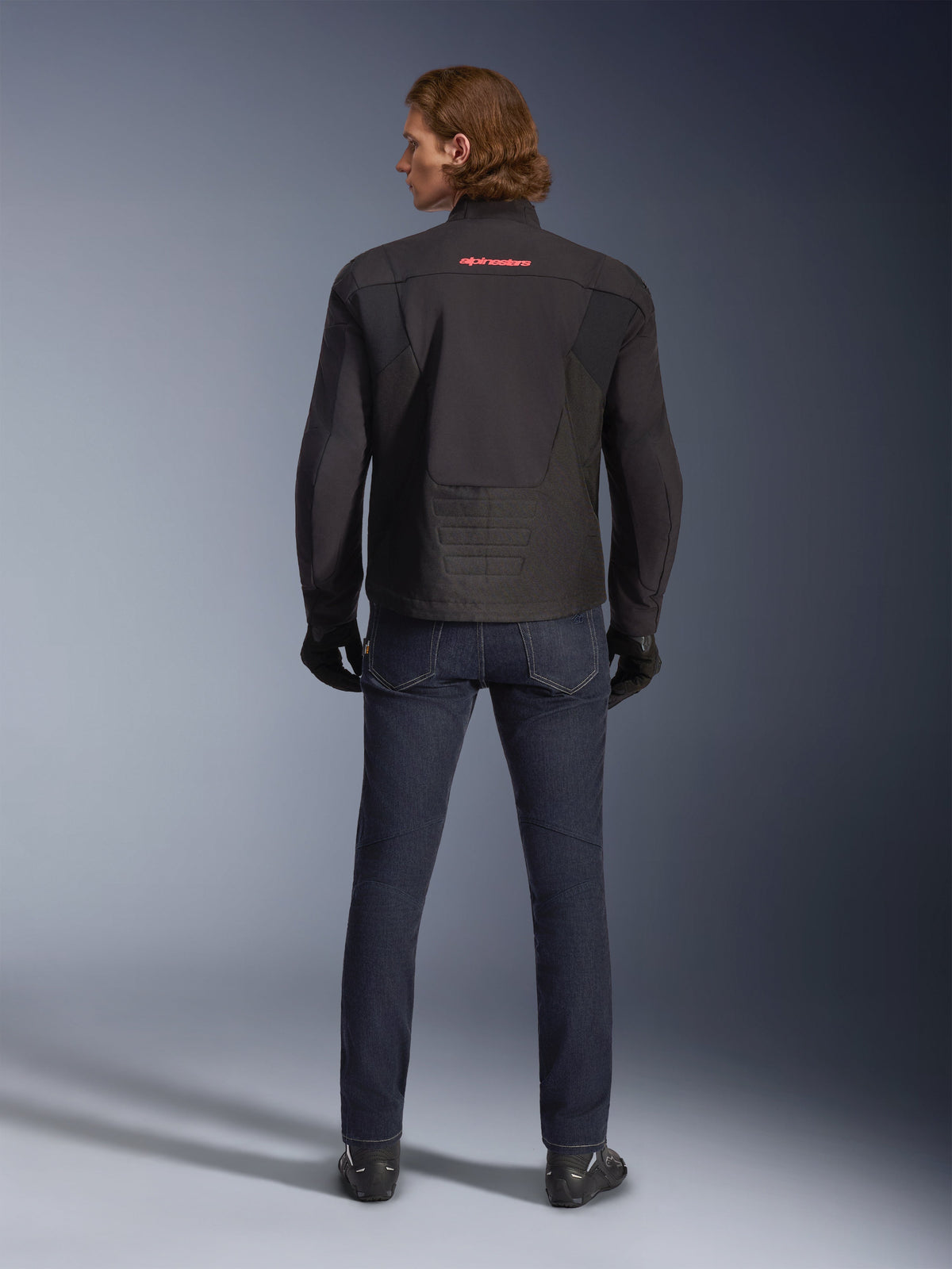 Alpinestars SMX Waterproof Jacket, svart, sedd bakifrån buren av en modell, med röd logotyp på övre ryggen, förlängt bakparti för täckning vid körning, stylad med mörkblå motorcykeljeans och svarta handskar.