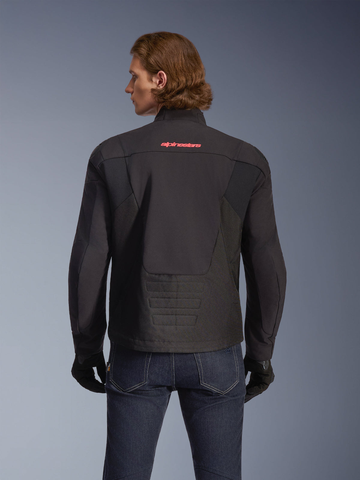 Alpinestars SMX Waterproof Jacket, svart med klarröd logotyp, vy bakifrån på en modell, bärs med mörkblå jeans och svarta handskar, textilmotorcykeljacka för väg- och sportkörning.