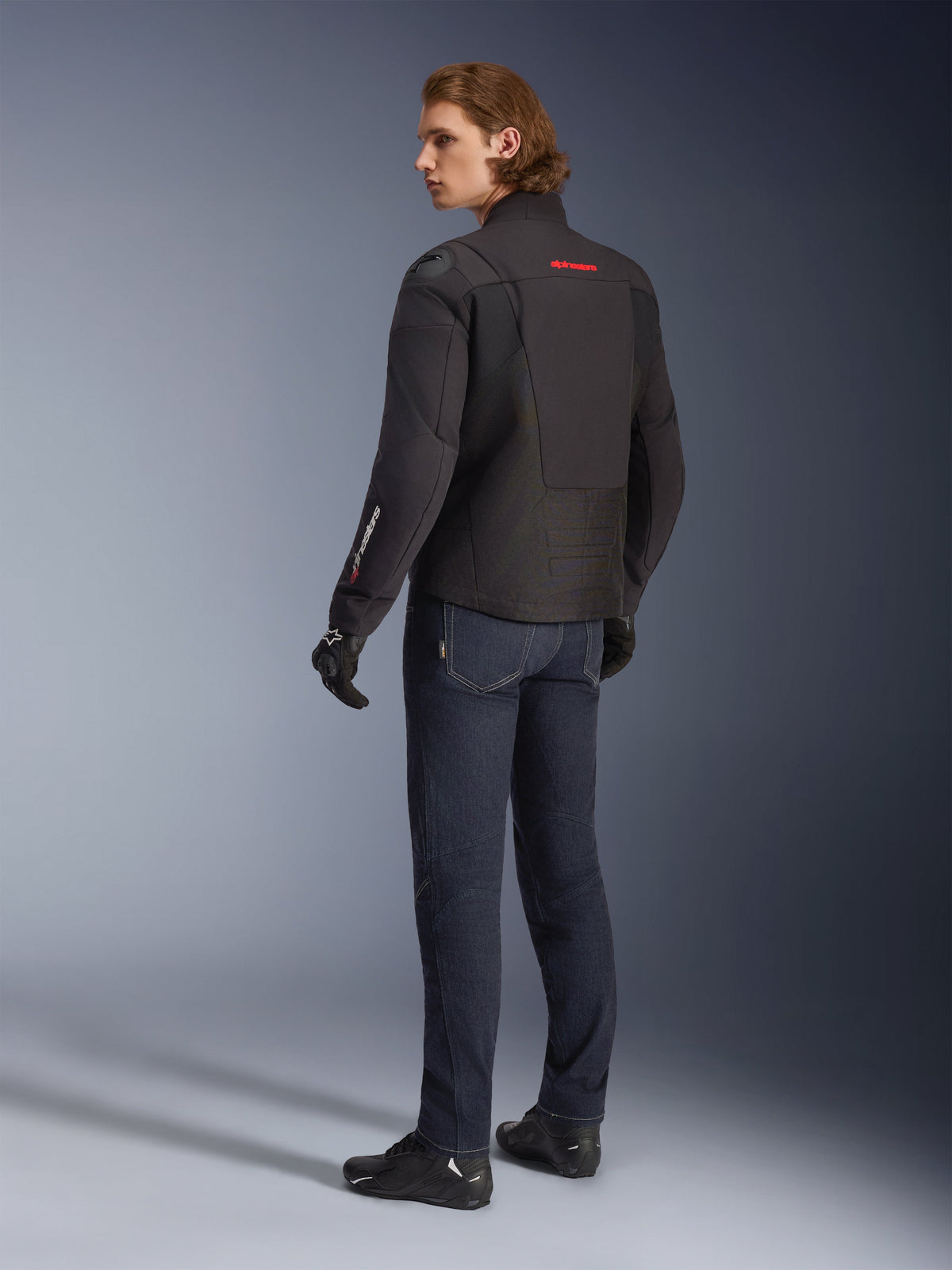 Alpinestars SMX Waterproof Jacket, svart motorcykeljacka med röda och vita detaljer, manlig förare sedd bakifrån iklädd mörkblå körjeans och svarta handskar, med externt axelskydd och förlängd bakre fåll för bättre täckning vid vägkörning