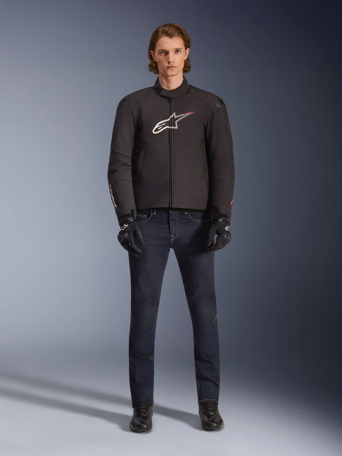 Modell som bär Alpinestars SMX Waterproof Jacket i svart med vit och röd logotyp, kombinerad med mörka mc-jeans i denim, svarta handskar och mc-stövlar, sportig motorcykeloutfit för alla väder