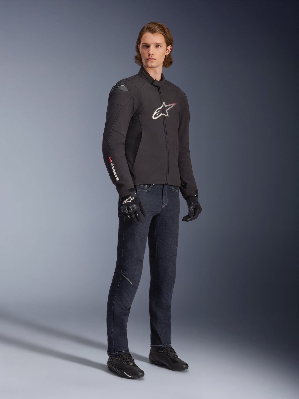 Manlig modell som bär Alpinestars SMX Waterproof Jacket, svart med vita och röda logotypdetaljer, stylad med mörkblå denimbyxor och svarta körhandskar, smidig motorcykeljacka i textil med sportig passform