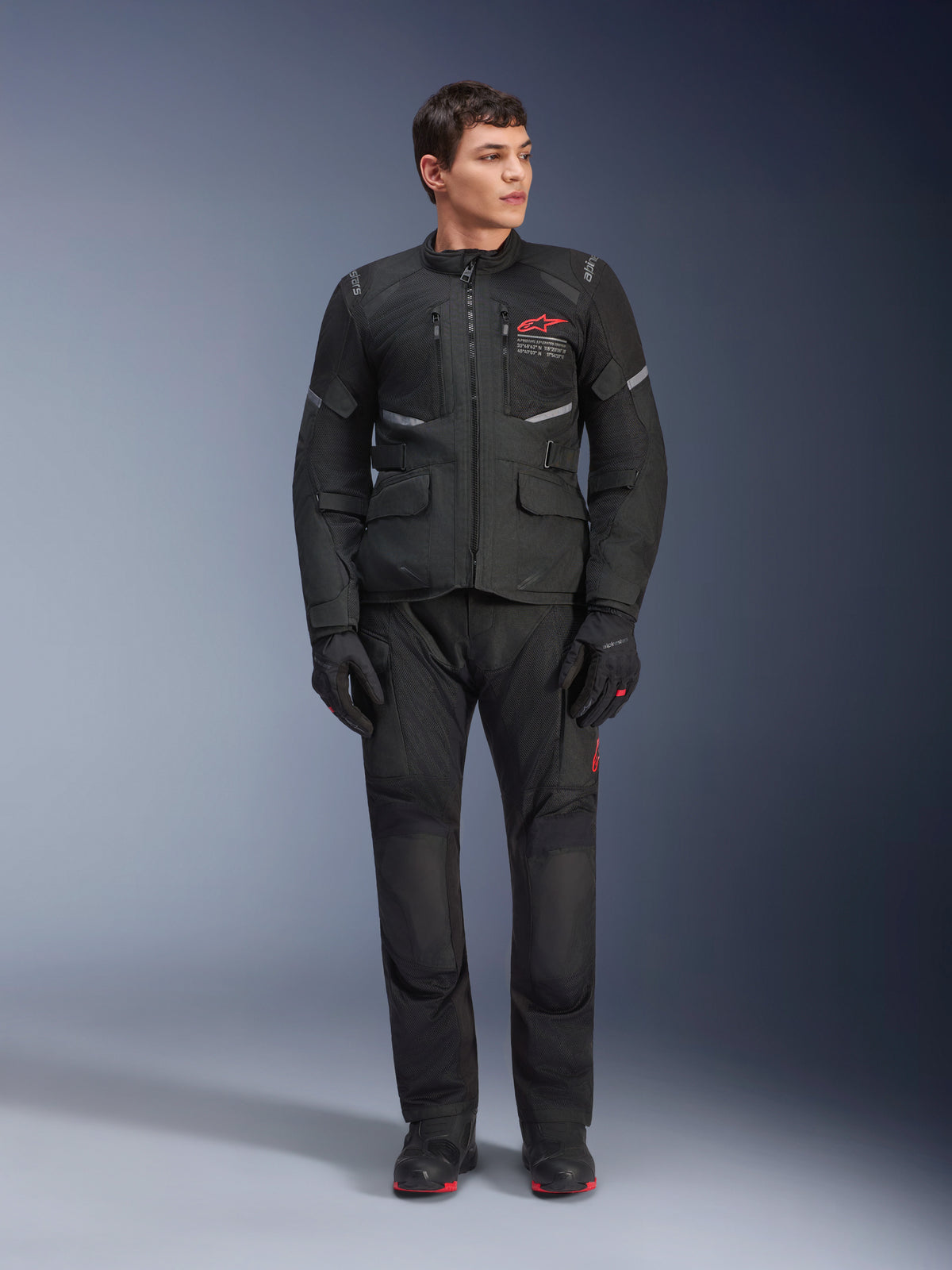 Manlig modell iklädd Alpinestars Andes Air Drystar®-jacka i svart, motorcykelutrustning för adventure touring med matchande byxor och handskar, röd logotyp på bröstet, nätventilation och funktionella fickor.