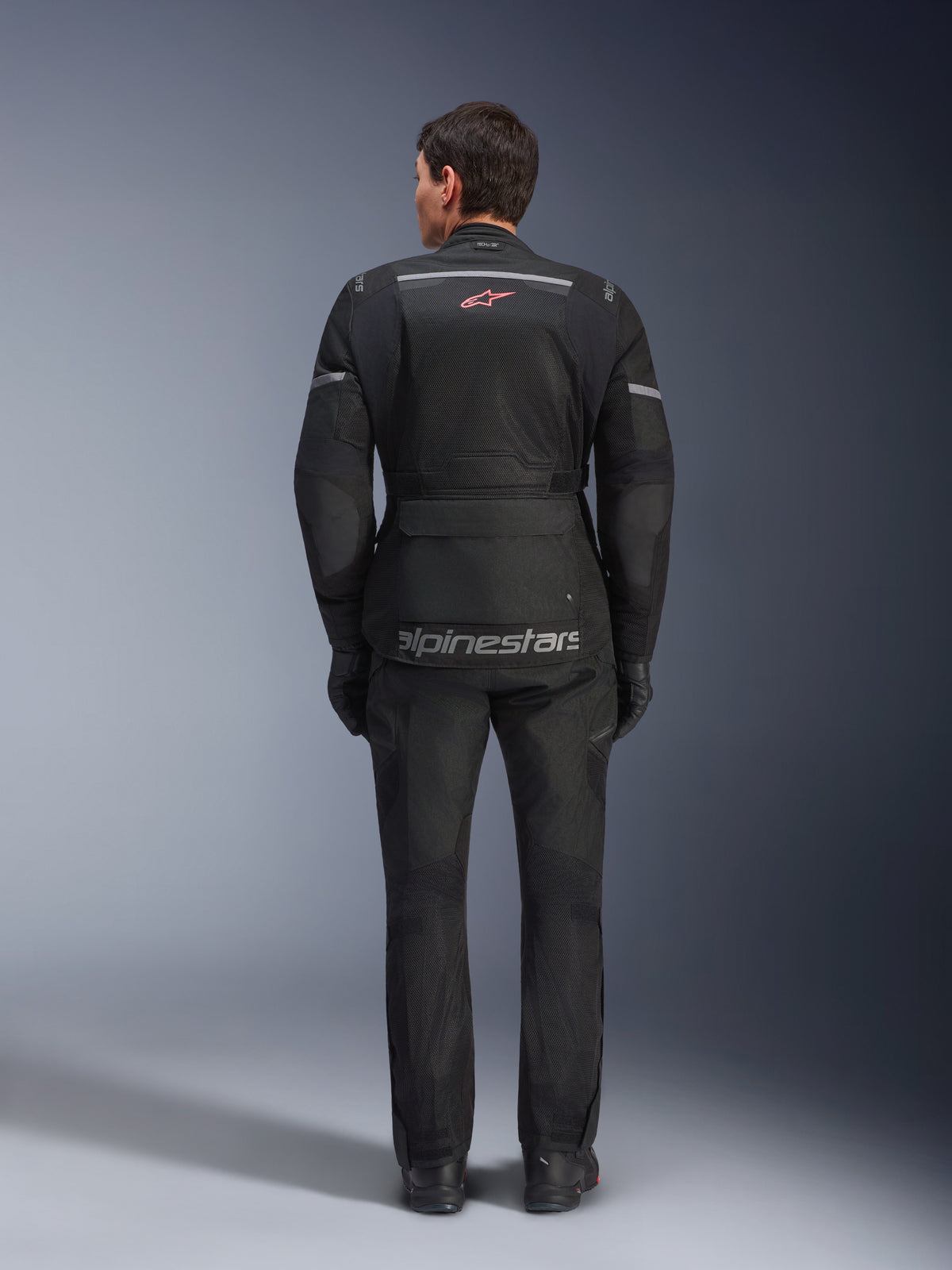 Vy bakifrån av en förare som bär Alpinestars Andes Air Drystar® Jacket i svart, med omfattande ventilationspaneler i mesh på ryggen, röd Alpinestars-logotyp och reflexdetaljer, kombinerad med matchande motorcykelbyxor och handskar för adventure touring.