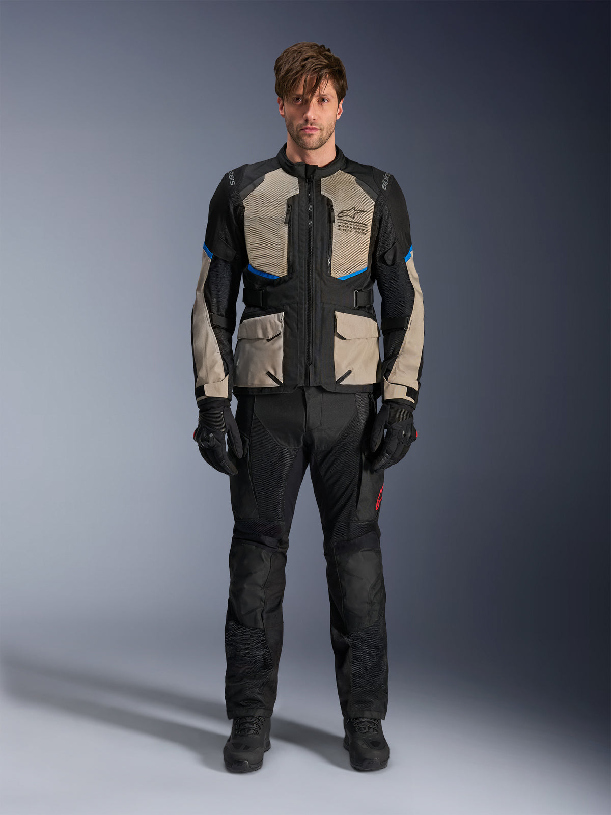 Modell som bär Alpinestars Andes Air Drystar®-jacka, touringjacka för motorcykel, svart, lagerbladsgrå och klarblå, matchad med svarta körbyxor och handskar, med omfattande ventilationspaneler i mesh på bröstet och ärmarna för adventure touring i varmt väder.