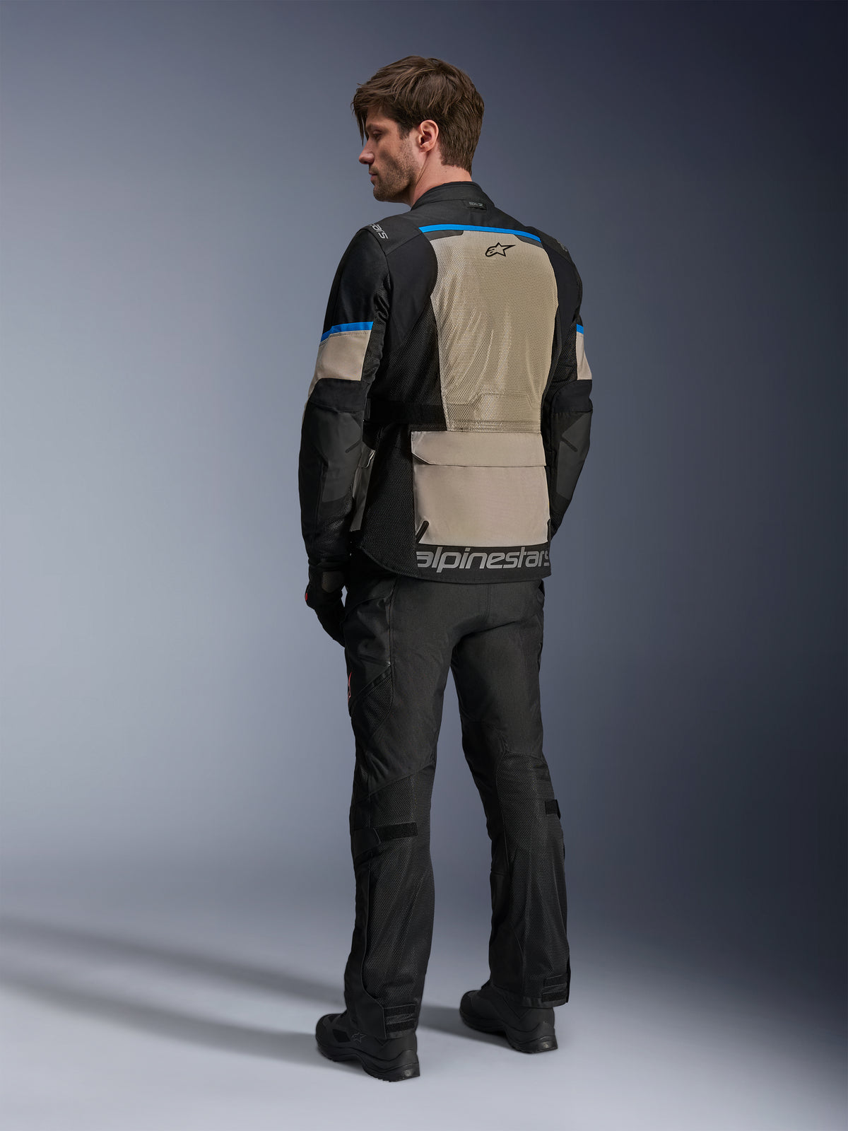 Alpinestars Andes Air Drystar®-jacka, svart, lagerbärsgrå och klarblå, vy bakifrån som visar en stor ventilationspanel i mesh på ryggen, logotyp på den nedre fållen, bärs med svarta motorcykelbyxor och stövlar.