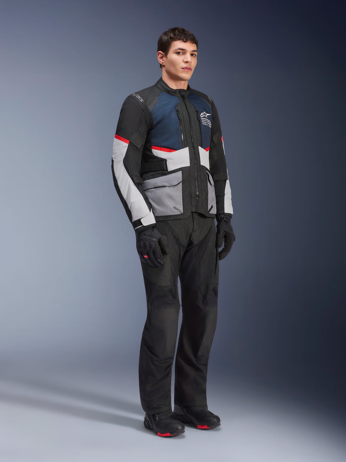Manlig modell bär Alpinestars Andes Air Drystar®-jacka i Deep Blue Black Ice Gray, kombinerat med svarta motorcykelbyxor, handskar och stövlar, med stora nätpaneler för ventilation, ljusgrå fickor och röda detaljer, designad för adventure touring