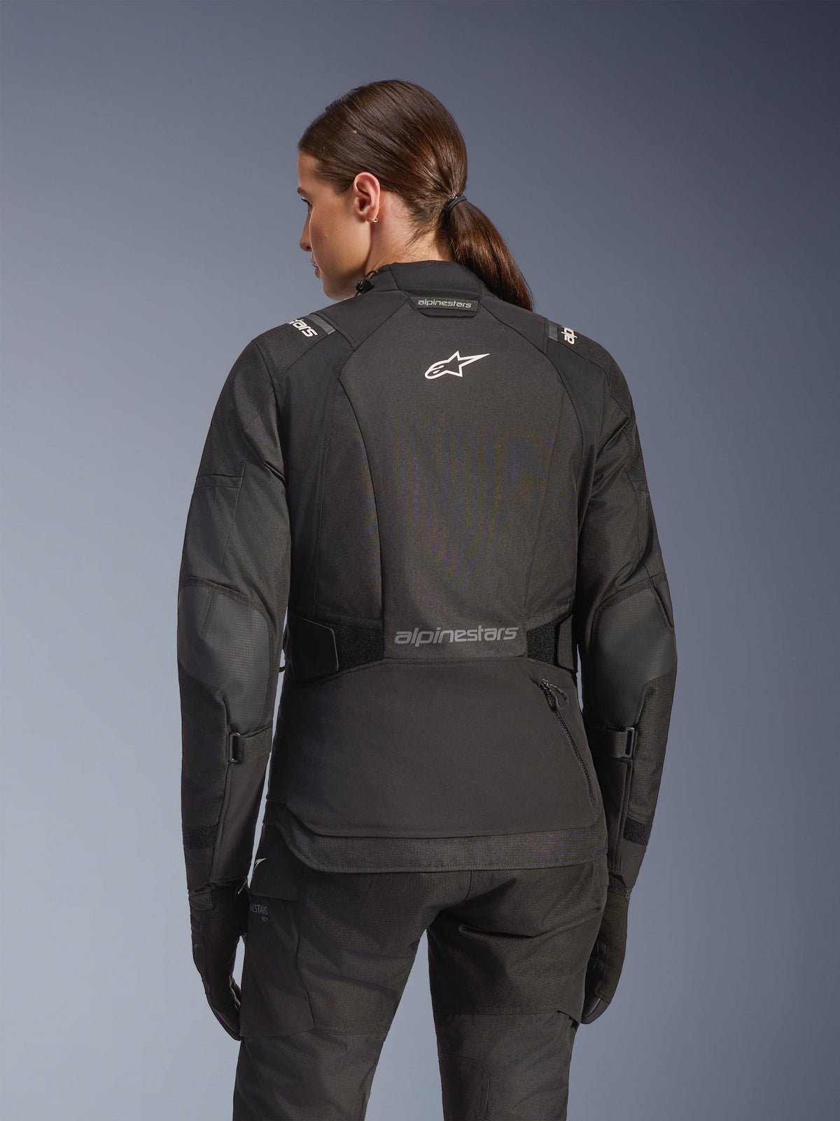 Kvinna bärande Alpinestars Stella Andes V4 Drystar-jacka i svart, ryggvy med vit logotyp på ryggen och förstärkta armbågspaneler, kombinerat med matchande svarta byxor och handskar, motorcykeloutfit för adventure touring