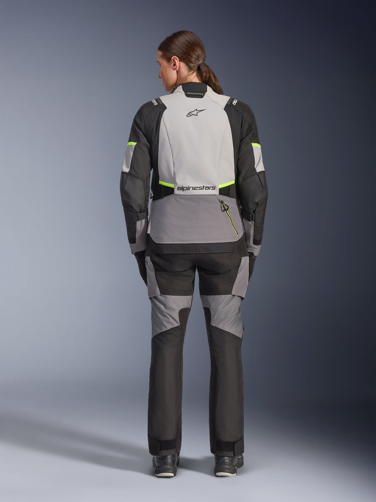 Kvinnlig förare sedd bakifrån i Alpinestars Stella Andes V4 Drystar®-jacka och matchande byxor, Ice Gray och Dark Gray med detaljer i Black och Yellow Fluo, adventure-touring motorcykelutrustning med hållbar textilkonstruktion och reflekterande logotyper