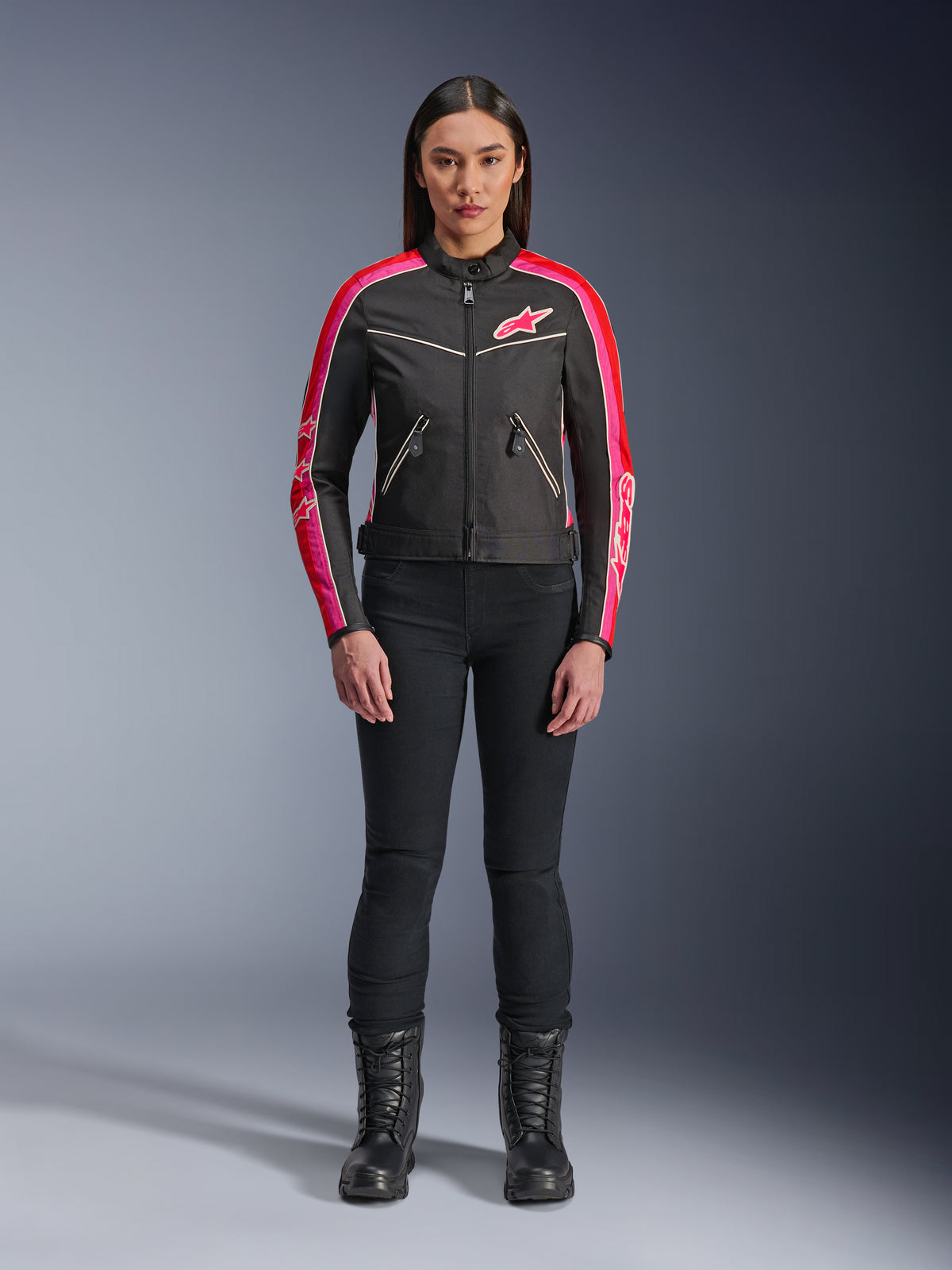 Kvinna iklädd Alpinestars Stella T-Dyno WR-jacka, svart med klarröda och fuchsiafärgade detaljer, textiljacka i vintagestil med rosa logotyp och ränder på ärmarna, bärs med svarta byxor och kängor med snörning, urban motorcykelutrustning.