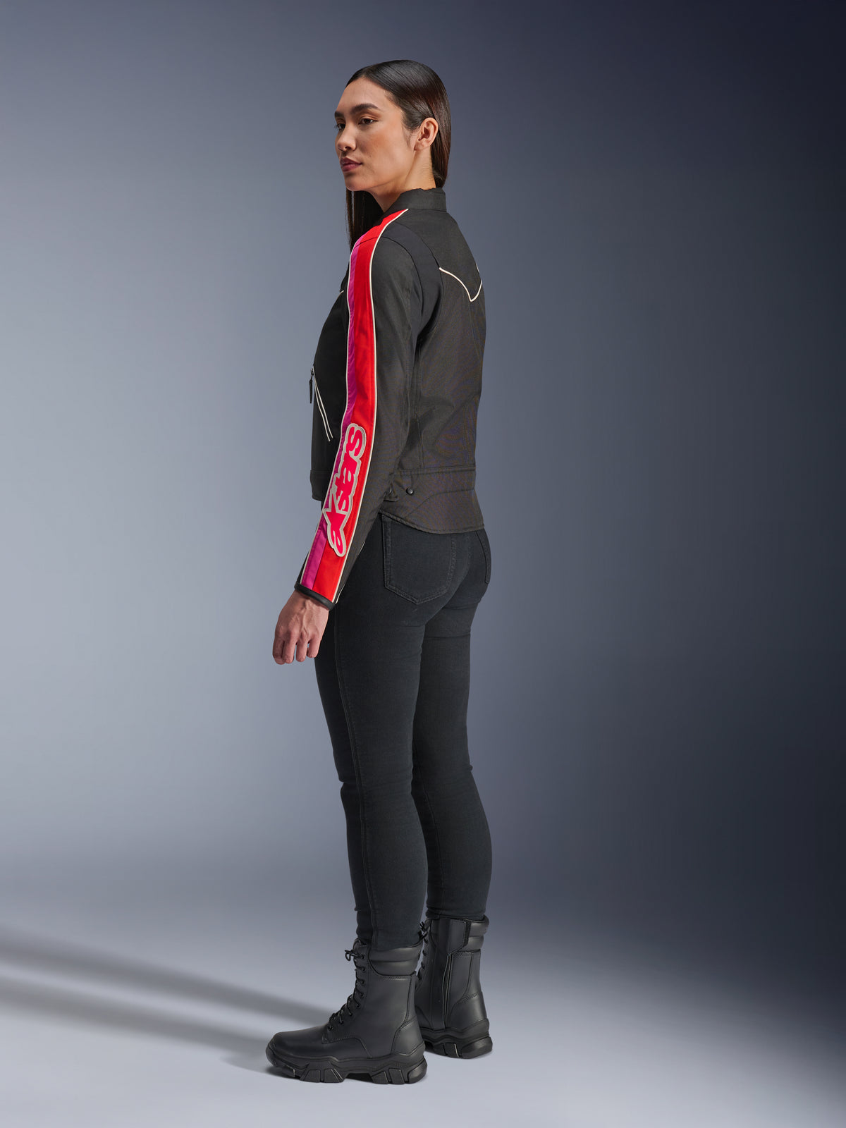Kvinnlig modell i Alpinestars Stella T-Dyno WR-jacka i Black Bright Red Fuchsia, med kraftfulla röda och rosa ränder och Alpinestars logotyp på ärmen, matchat med svarta motorcykelbyxor och svarta stövlar i en studiomiljö.