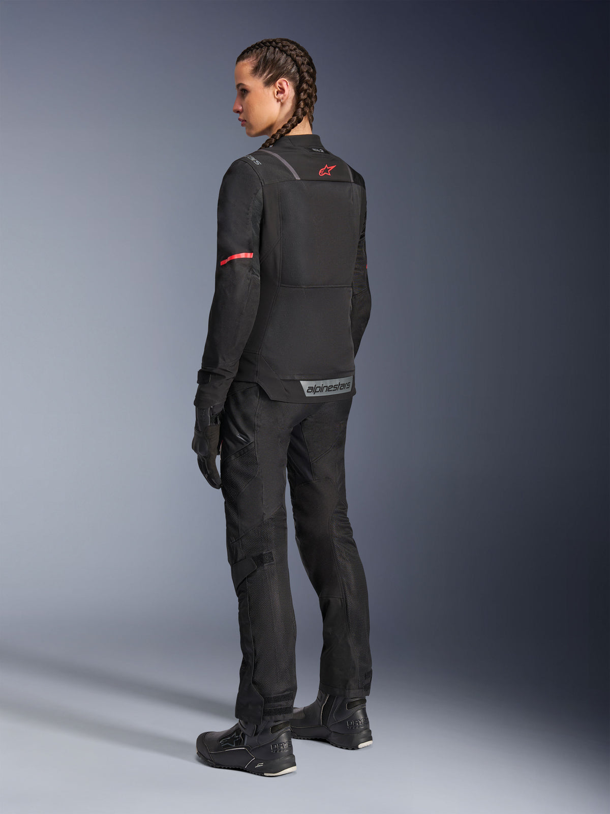 Kvinnlig förare i svarta Alpinestars Stella Andes Air Drystar®-byxor och matchande Stella ST-2 Air-jacka, vy bakifrån som visar teknisk textilkonstruktion och meshpaneler, röd stjärnlogotyp på jackans rygg, kombinerat med svarta mc-stövlar och handskar.
