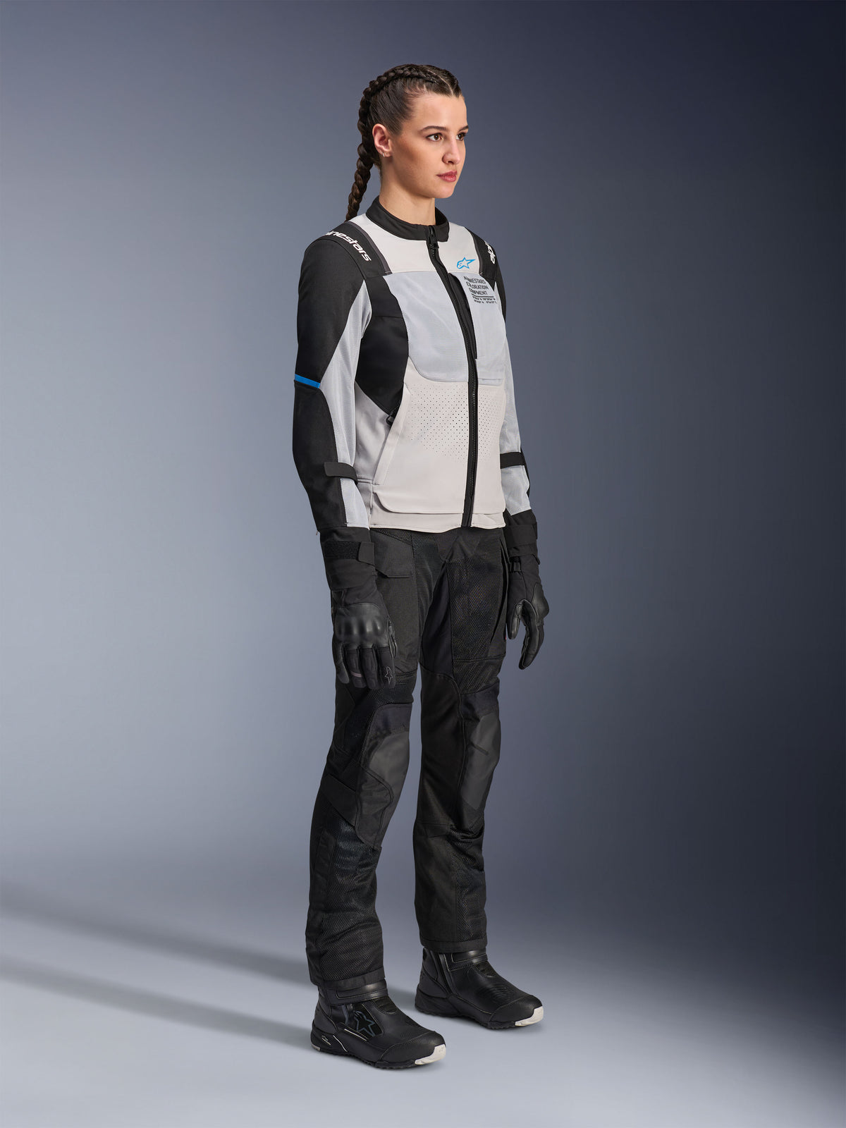 Modell som bär Alpinestars Stella ST-2 Air Jacket i Ice Gray och svart, teknisk motorcykeljacka för dam med meshpaneler och softshell-konstruktion, kombinerad med svarta adventure touring-byxor och stövlar, med blå detaljer och en modern design för MC-åkning