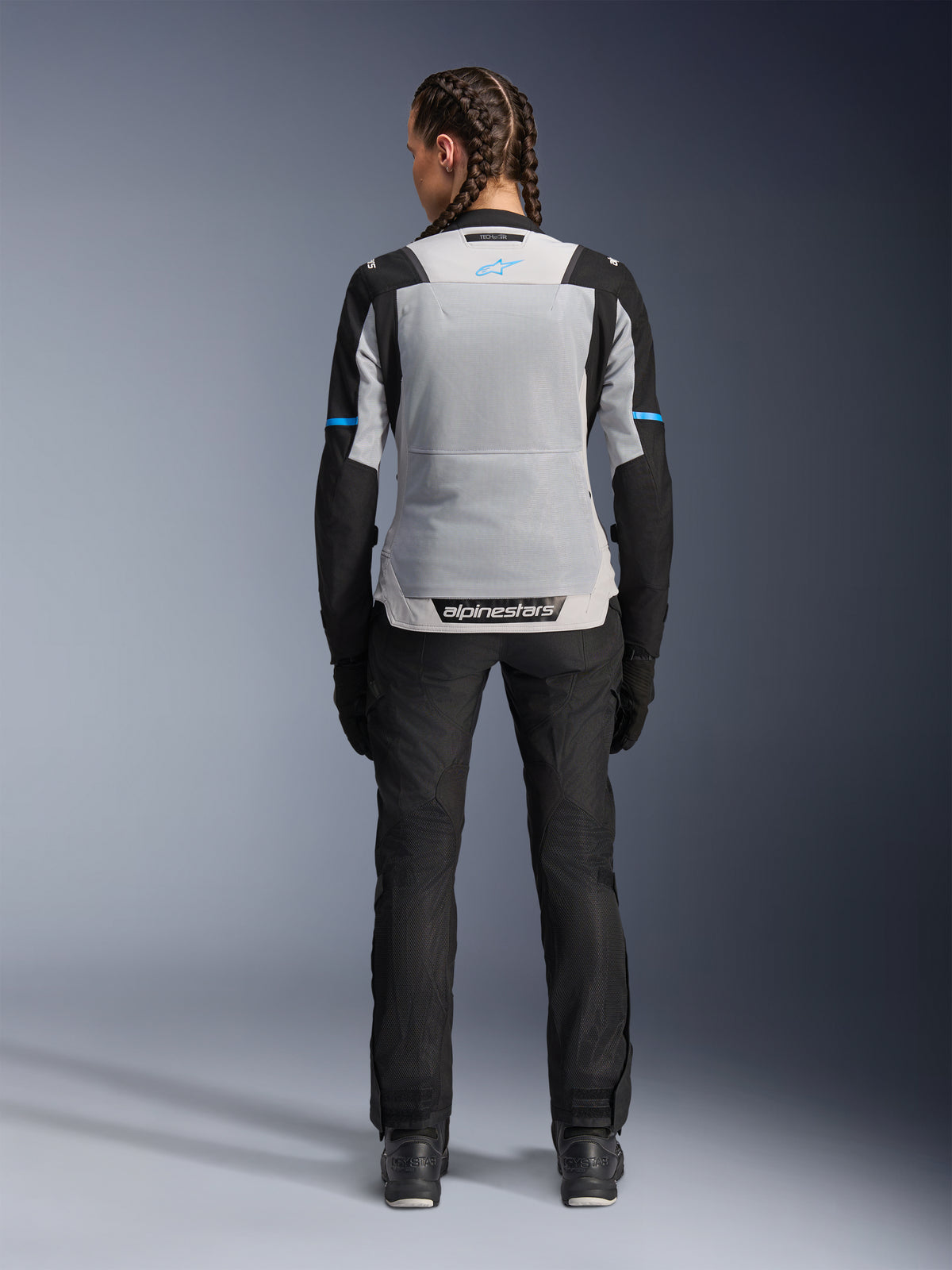 Alpinestars Stella ST-2 Air-jacka, Ice Gray Black, vy bakifrån på kvinnlig modell, med en stor ljusgrå ryggpanel i mesh, svarta ärmar med blå detaljer, Alpinestars-logotyp på nedre delen av ryggen, matchad med svarta tekniska MC-byxor och stövlar, sommarutrustning för motorcykel.