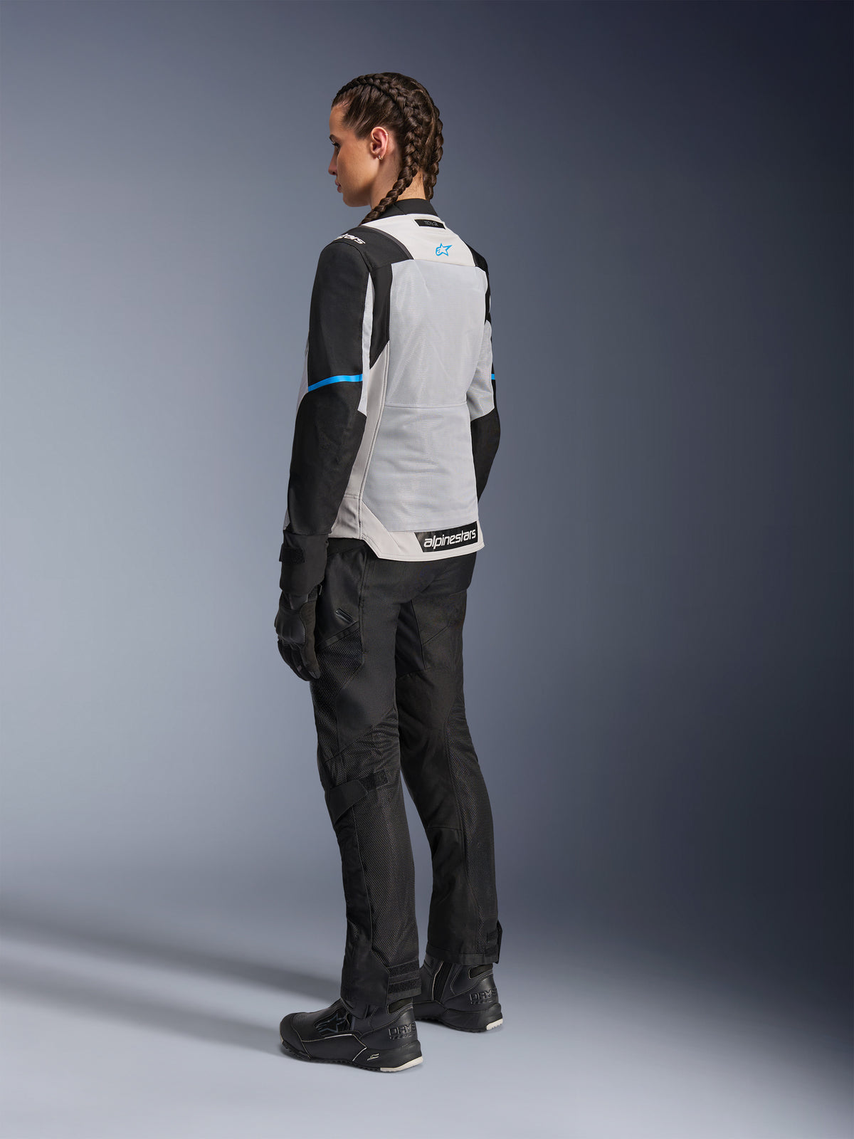 Kvinnlig förare klädd i Alpinestars Stella ST-2 Air-jacka i isgrå och svart, trekvartsvy bakifrån som framhäver den stora ljusgrå meshpanelen på ryggen, svarta ärmar med en blå accentrand och en blå Alpinestars-logotyp, matchat med svarta mc-byxor, handskar och stövlar för en komplett adventure touring-outfit