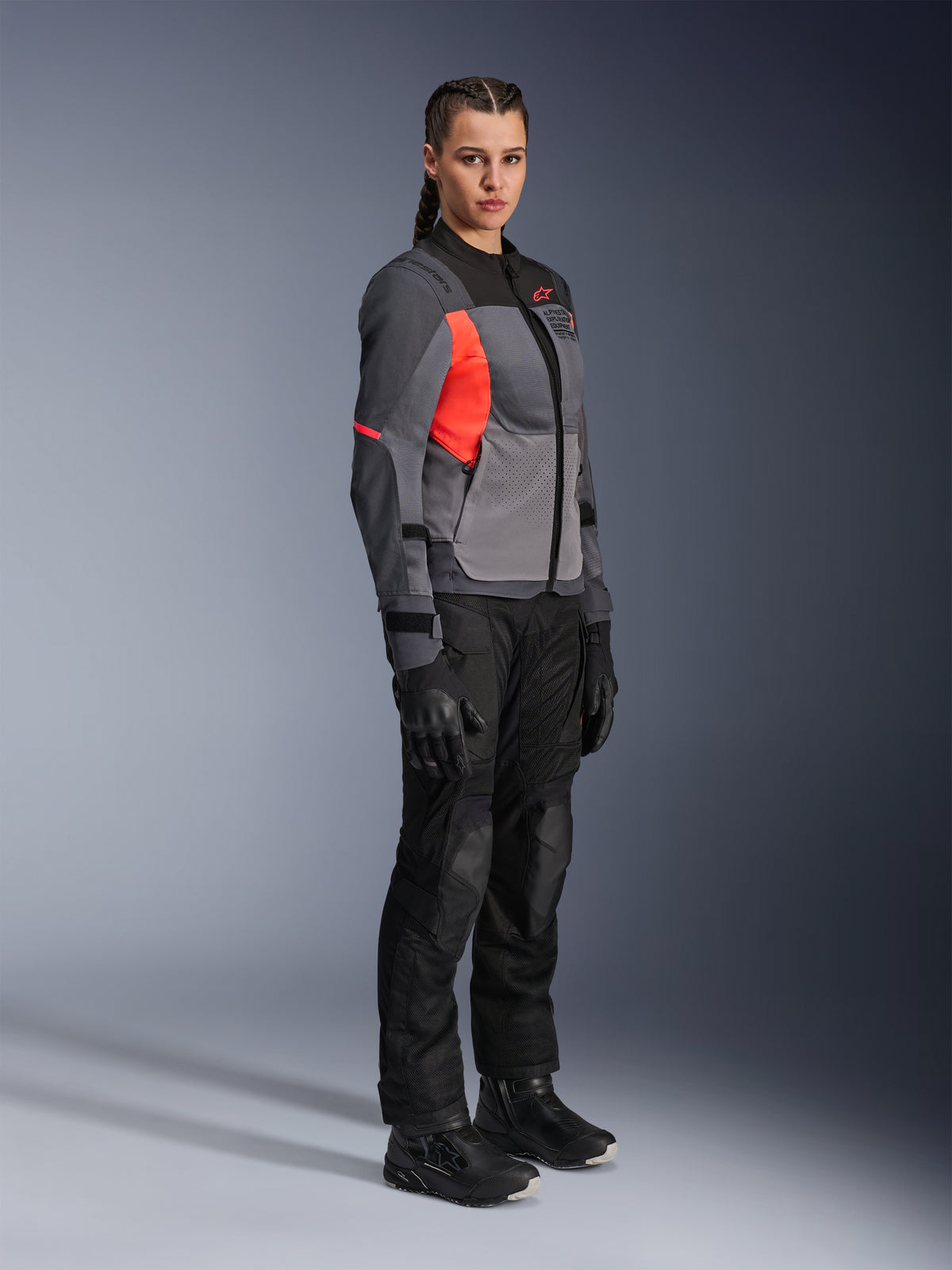 Kvinnlig förare som bär Alpinestars Stella ST-2 Air Jacket i Deep Gray, Dark Gray och Fluo Coral, kombinerat med svarta motorcykelbyxor, handskar och stövlar, med en modern touring-design med meshpaneler och reflekterande detaljer.