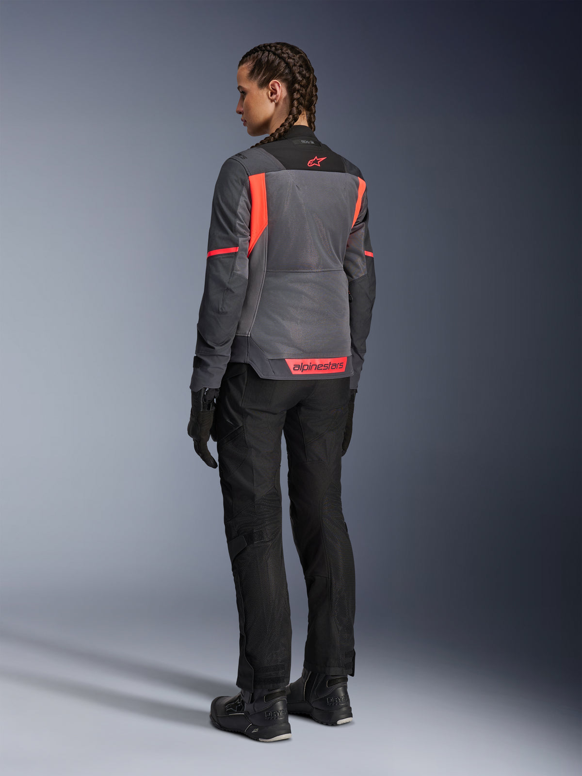 Kvinna som bär Alpinestars Stella St-2 Air-jacka, motorcykeljacka för touring, Deep Gray och Dark Gray med detaljer i Fluo Coral, matchad med svarta körbyxor och handskar, vy bakifrån som visar stora meshpaneler och reflekterande märkning i korallfärg, designad för sommarens mc-äventyr