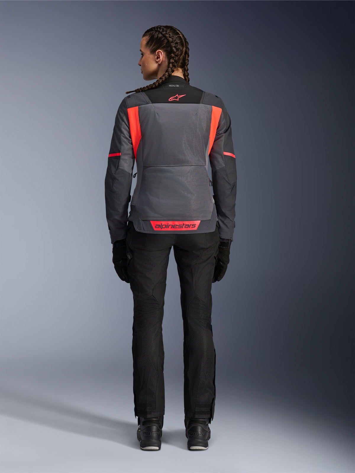 Ryggvy av en modell som bär Alpinestars Stella St-2 Air-jacka för dam i Deep Gray och Dark Gray med Fluo Coral-detaljer, med stora ventilationspaneler i mesh på ryggen och reflekterande logotypdetaljer, matchad med svarta motorcykelbyxor och handskar.