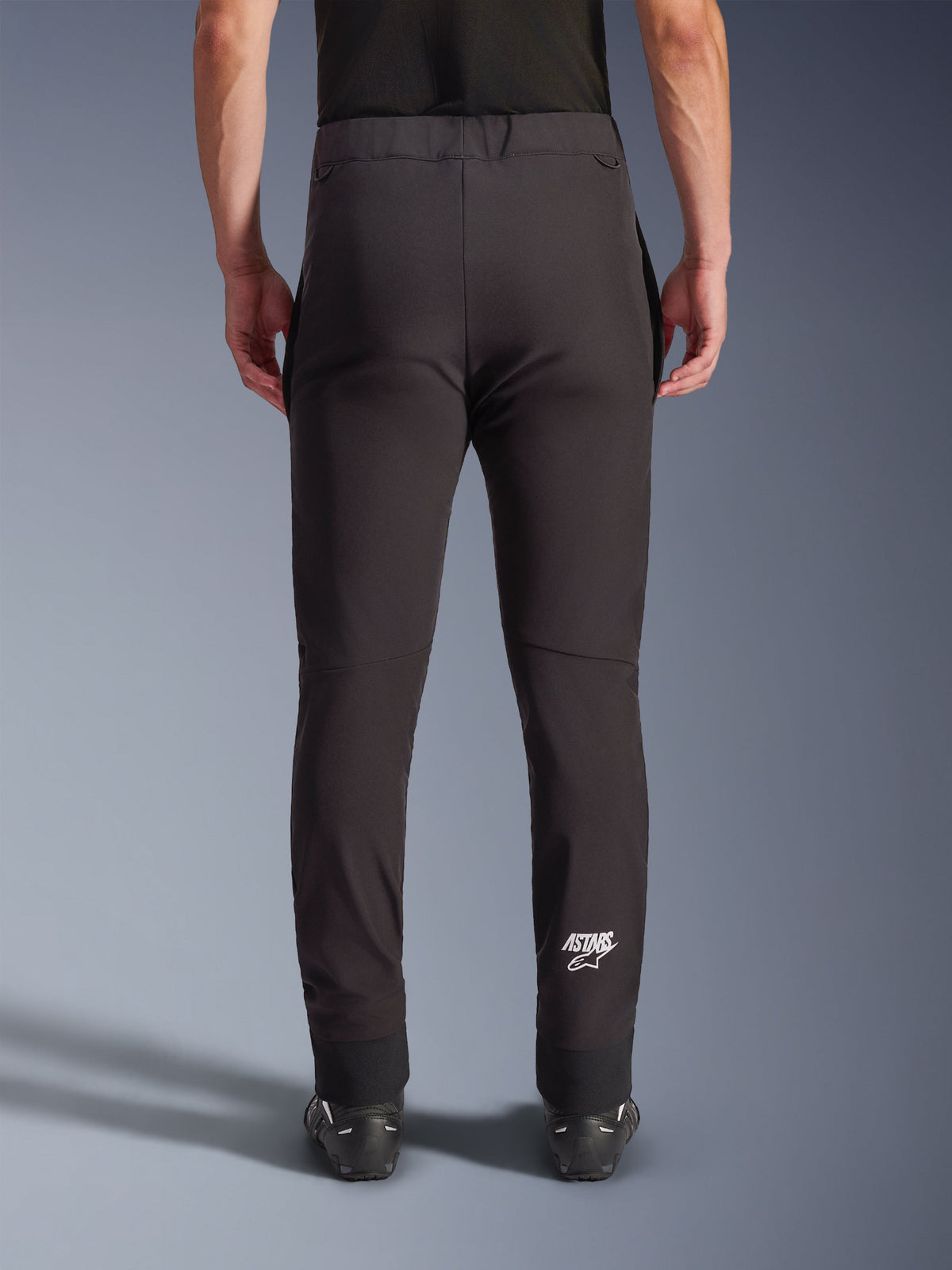 Alpinestars Aeroshell Pants, Black softshell motorcycle pants, back view showing slim fit and pre-curved knees, white Astars logo on the lower right calf, elasticated lower hem, crossover design for road riding