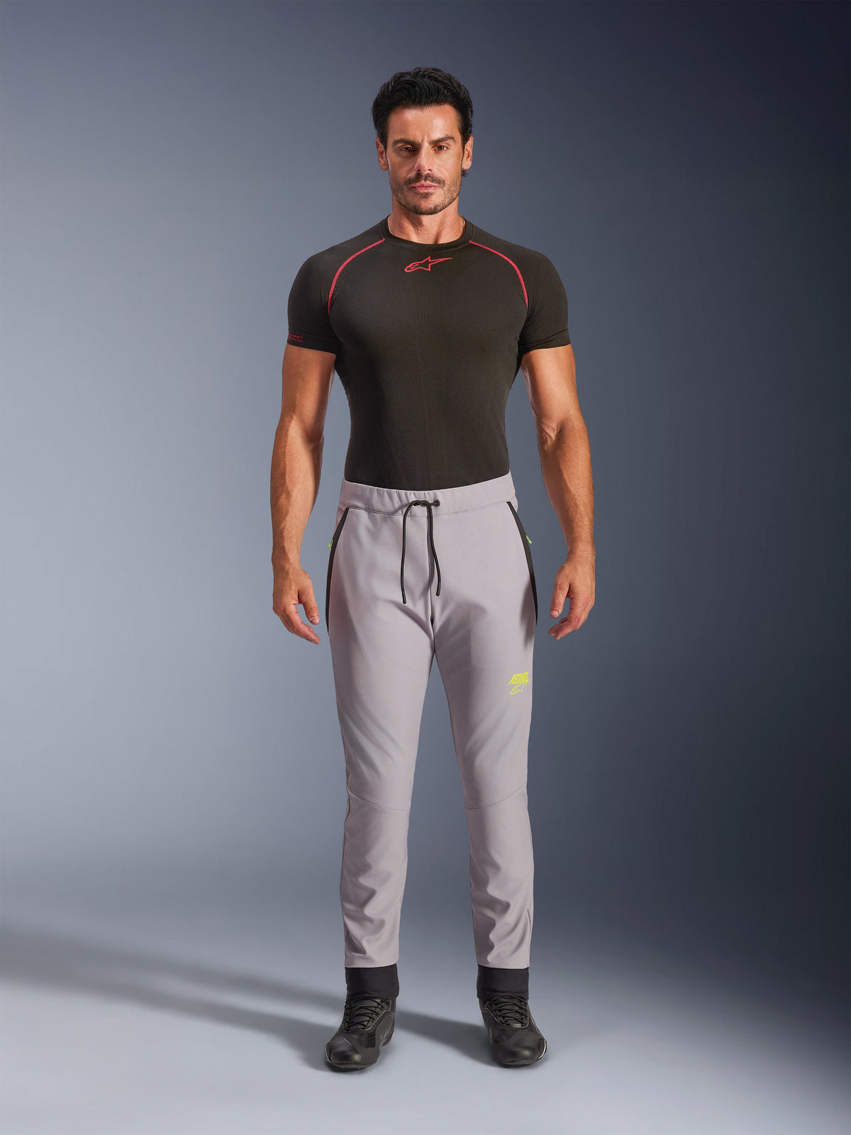 Manlig modell i Alpinestars Aeroshell-byxor i Mid Gray och Yellow Fluo, crossover sportiga softshellbyxor med elastisk midja med dragsko och fickor med dragkedja, kombinerat med en svart kortärmad funktionströja, modern atletisk design för landsvägs- och vardagskörning