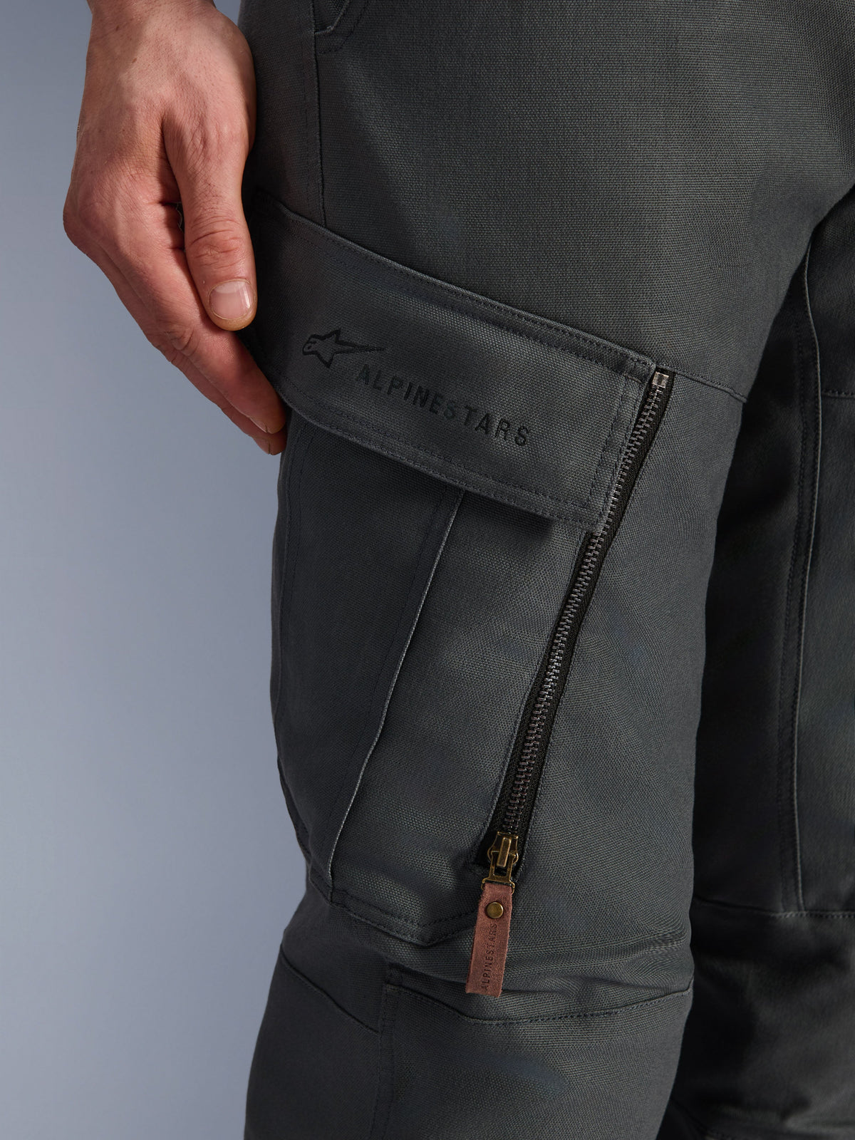 Alpinestars Borrego Drystar®-byxor, antracitgrå, närbild på lårets cargoficka med broderad logotyp, strukturerat vaxat canvasmaterial, vertikal ventilationsdragkedja med brun dragflik i läder