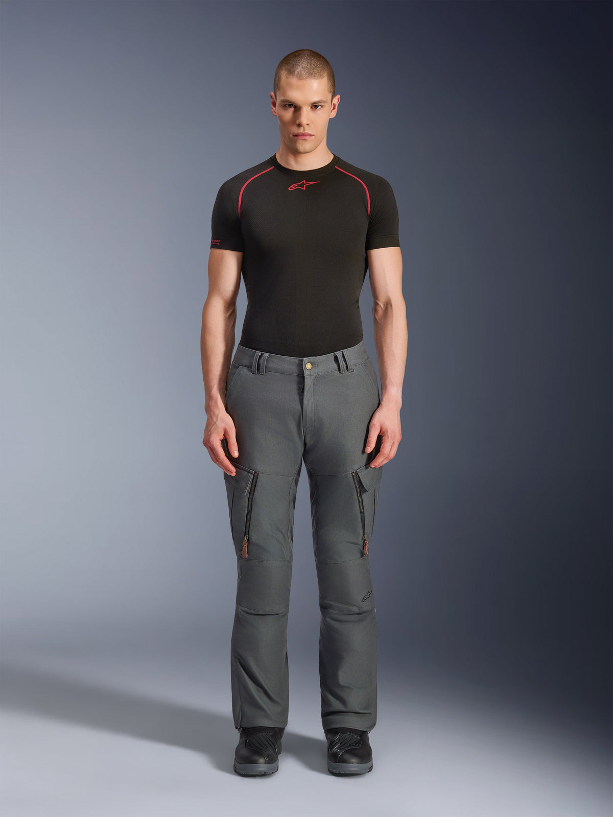 Alpinestars Borrego Drystar®-byxor, grå äventyrsmotorcykelbyxor, bärs av en modell med en svart och röd teknisk t-shirt och svarta stövlar, med cargofickor med vertikala dragkedjor, robust vaxad canvas-look, hållbar design för äventyrstouring och landsvägskörning