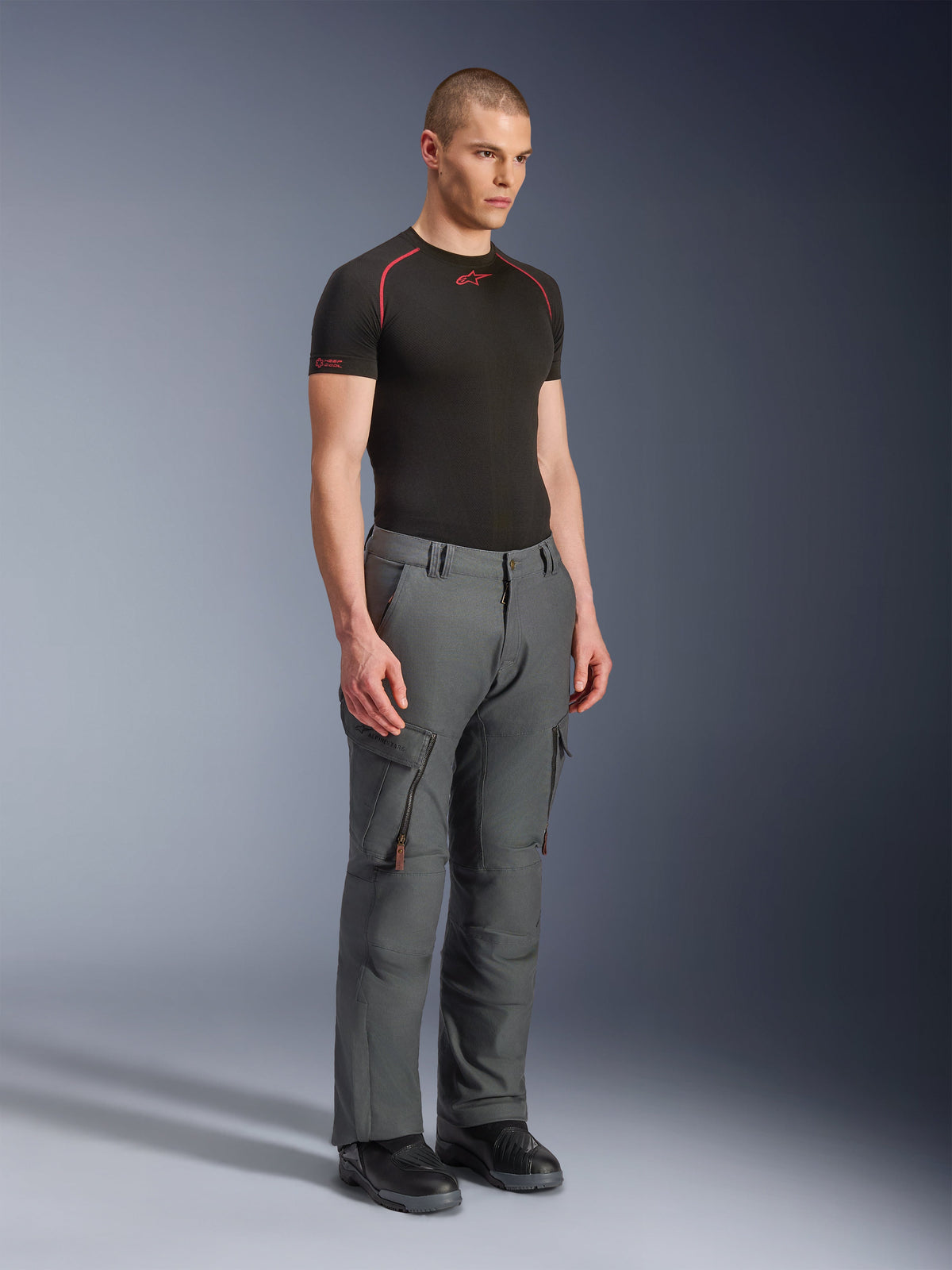 Manlig modell bär Alpinestars Borrego Drystar®-byxor, grå antracitfärgade äventyrsmotorcykelbyxor, arbetsklädesinspirerad design med cargofickor och dragkedjeförsedda ventiler, stylade med ett svart kortärmat baslager och svarta stövlar