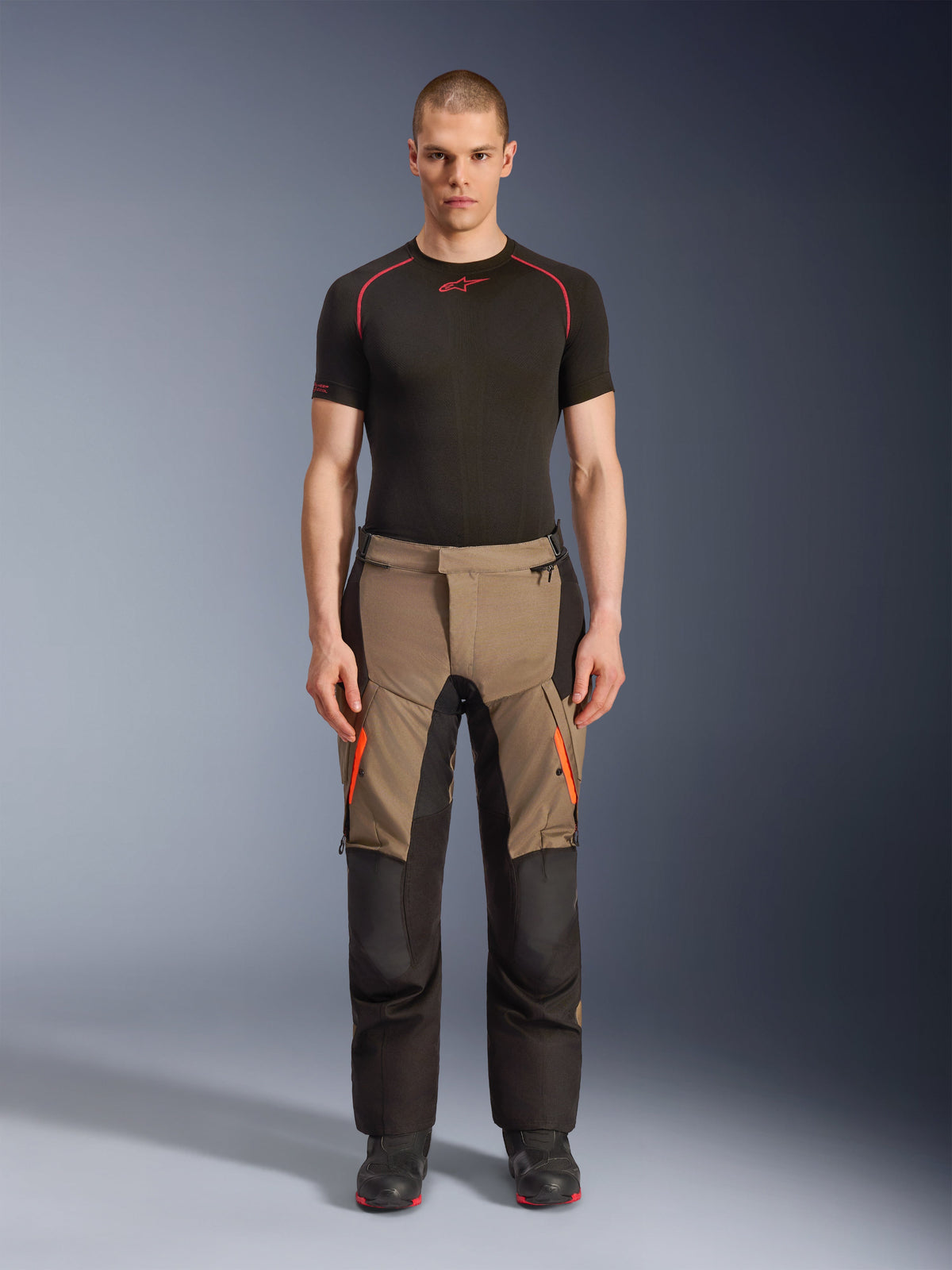 Manlig modell som bär Alpinestars Andes V4 Drystar® Byxor i Walnut Black, äventyrsmotorcykelbyxor kombinerade med ett svart underställ och stövlar, med orangea dragkedjedetaljer på låren och förstärkta svarta paneler för hållbarhet i all terräng.