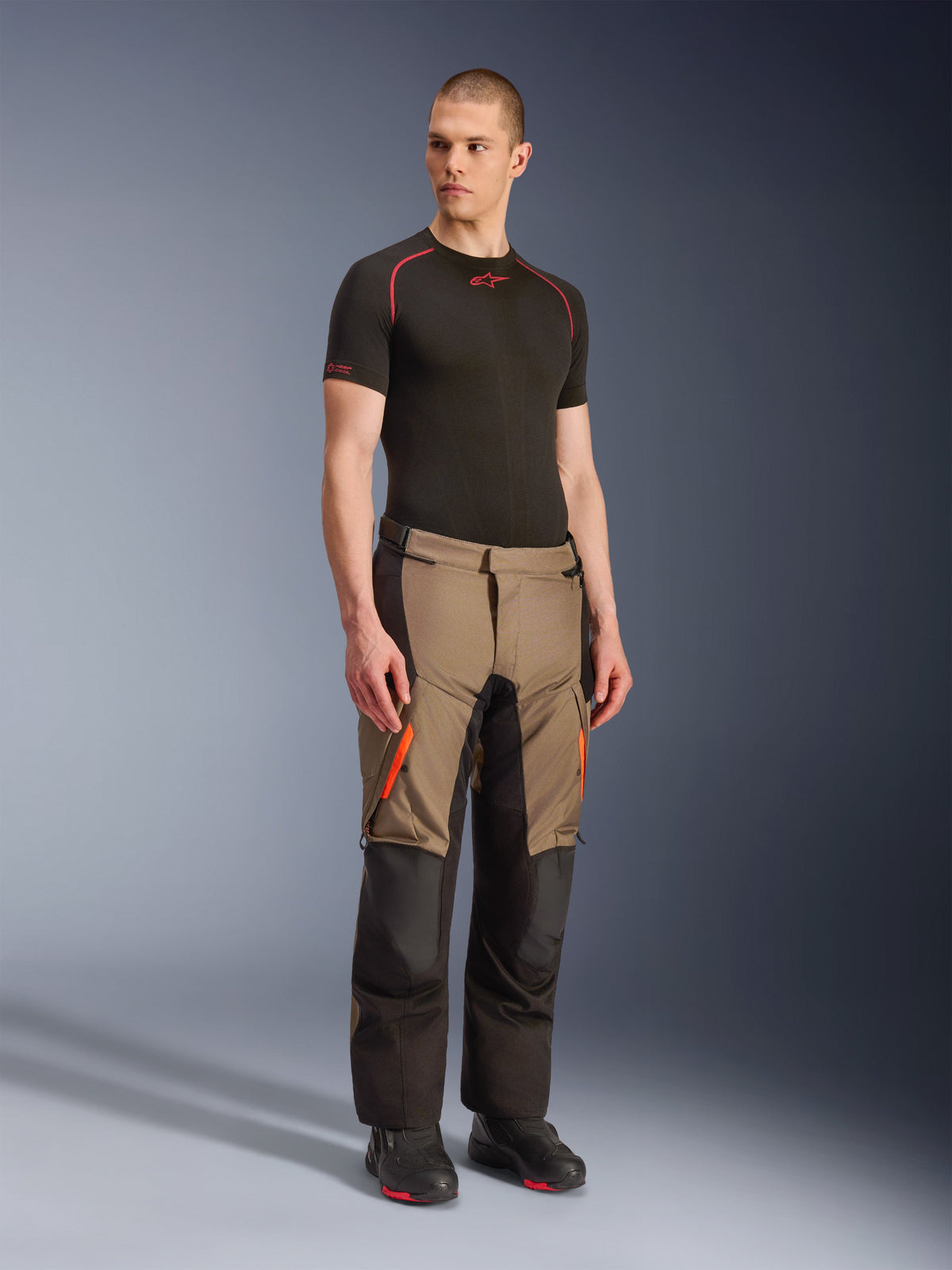 Alpinestars Andes V4 Drystar® byxor, äventyrs- och touringbyxor för motorcykel, Walnut Black med orangea detaljer, bärs av modell med svart kortärmat underställ och svarta stövlar, förstärkta textilpaneler och cargofickor, robust design för körning i alla väder