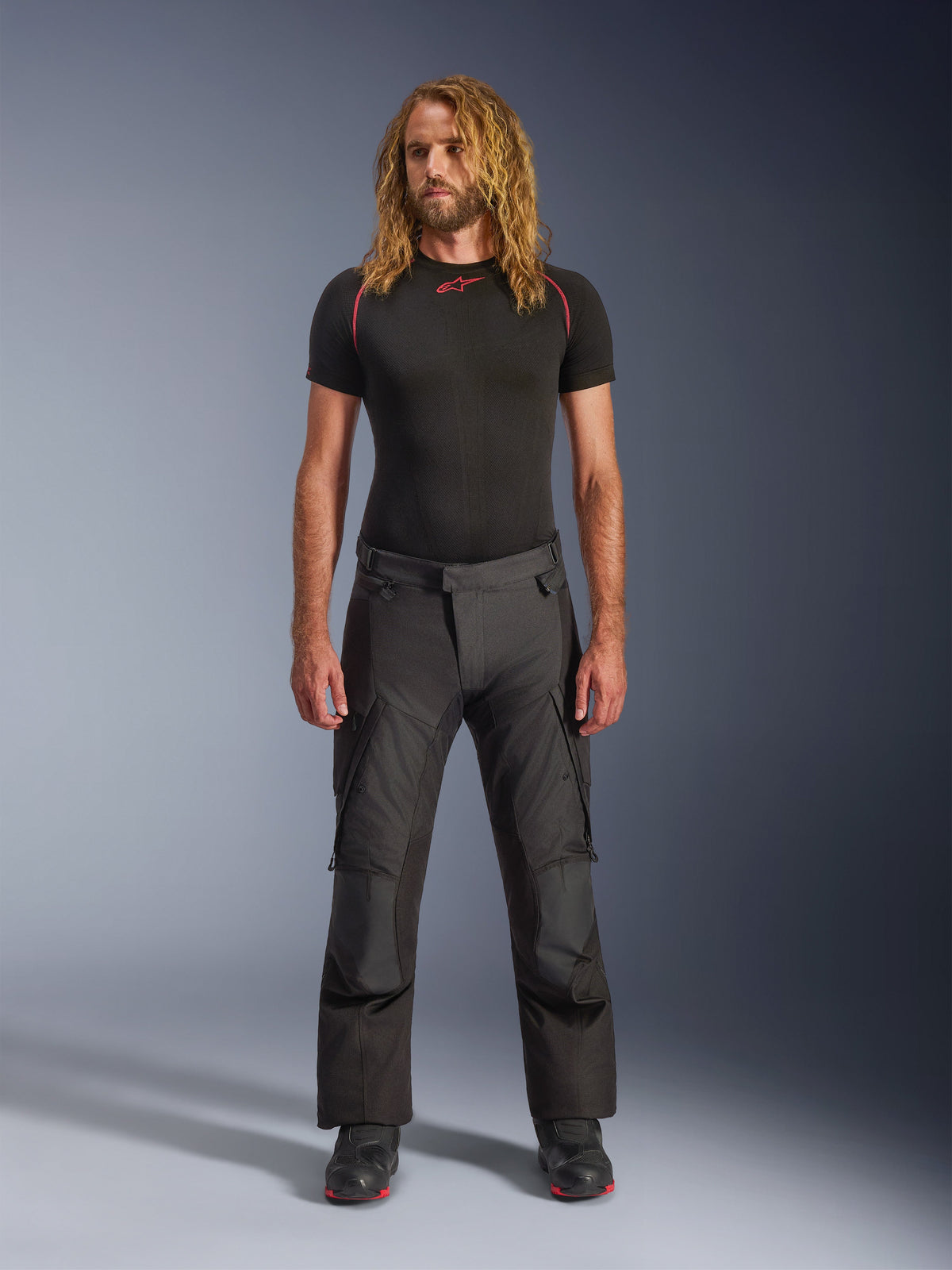 Manlig modell med långt hår iklädd Alpinestars Andes V4 Drystar®-byxor i svart, äventyrstouring-motorcykelbyxor med förstärkta knäpaneler och cargofickor, stylade med ett svart kortärmat baslager och körstövlar.