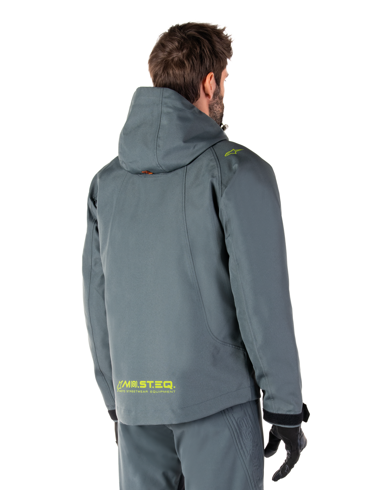 Alpinestars Morush Waterproof Parka i Urban Gray, sedd bakifrån, med gul MO.ST.EQ.-logotyp på nedre delen av ryggen och på axeln, minimalistisk design med huva, bärs med matchande grå mc-byxor och svarta handskar, urban mc-streetwear.
